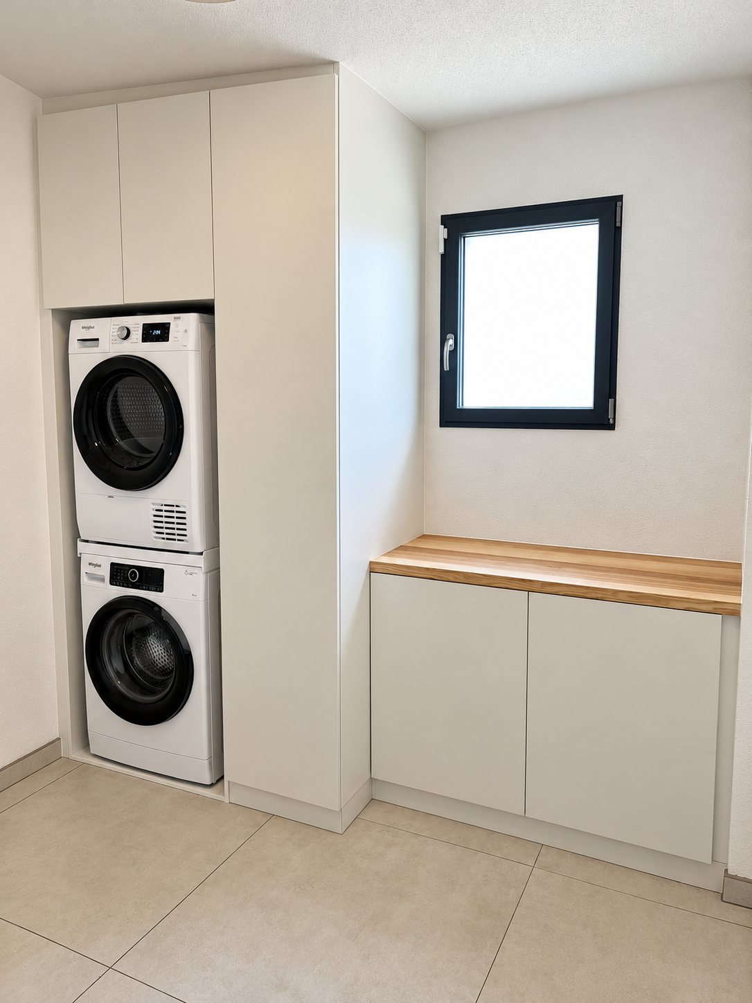 Buanderie aménagée avec une colonne de placards blancs abritant un lave-linge et un sèche-linge superposés, et un meuble bas avec plan de travail en bois sous une petite fenêtre.