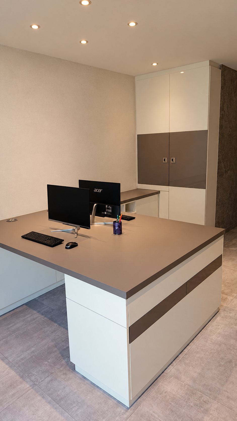 Un bureau d'angle blanc et taupe équipé de deux écrans d'ordinateur, installé dans une pièce lumineuse avec un grand placard assorti.