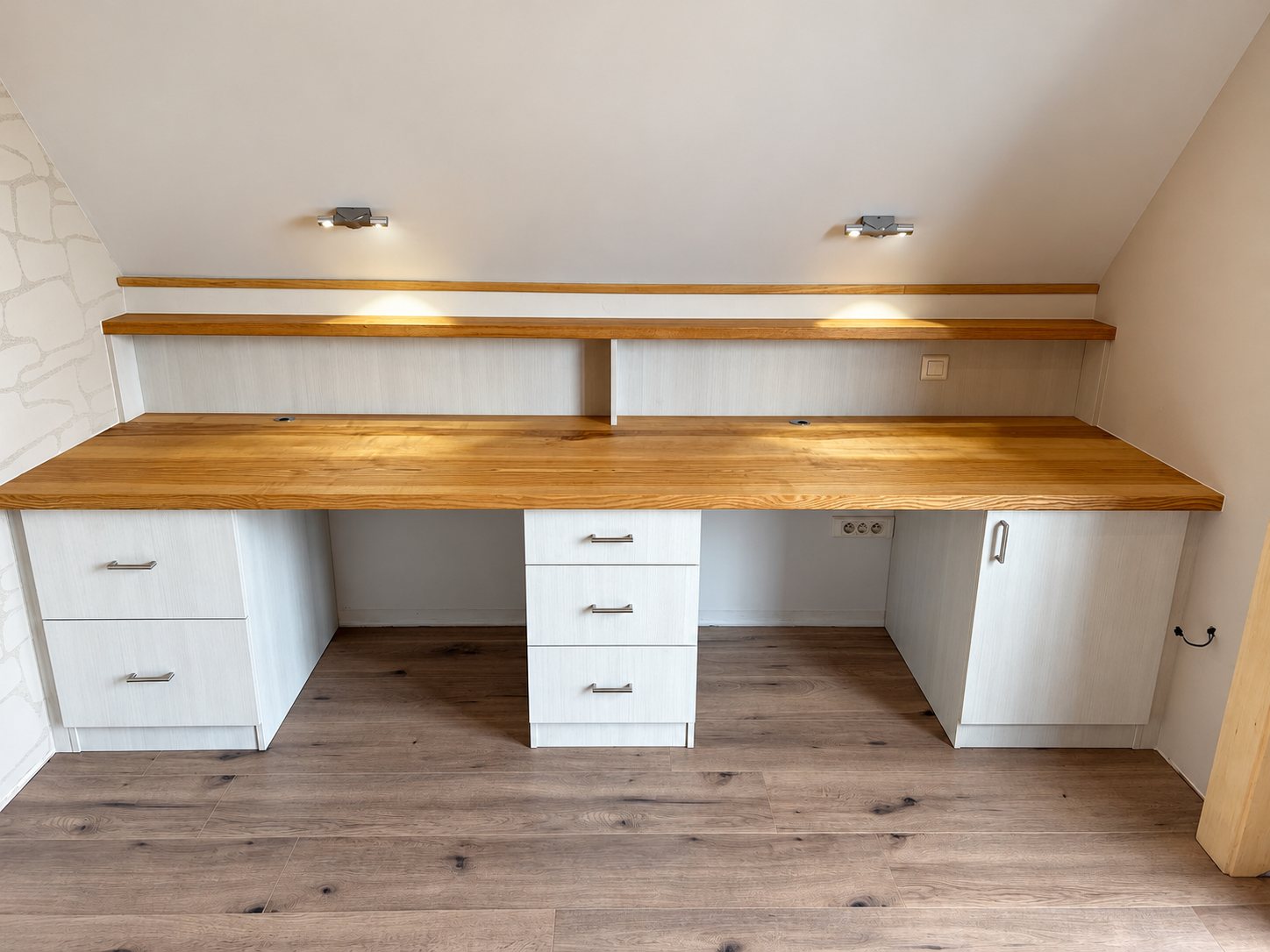 Un long bureau sur mesure avec plateau en bois massif, tiroirs et placard blancs et une étagère avec spots, installé sous un plafond mansardé.
