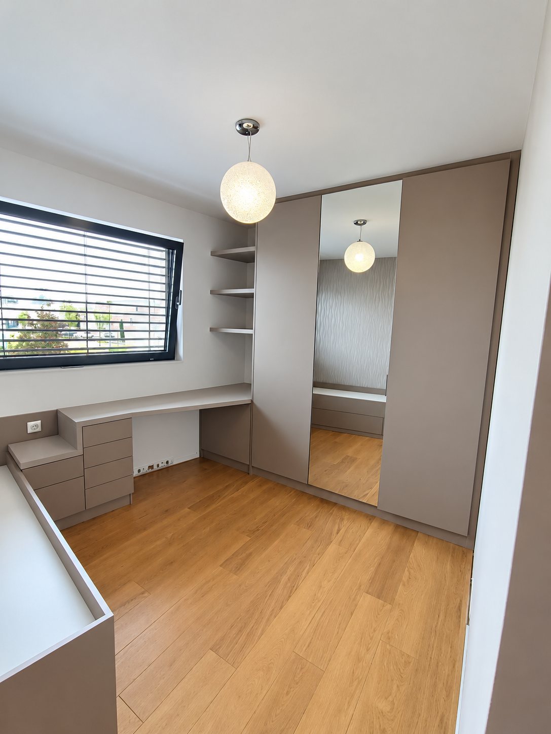 Chambre moderne avec dressing gris taupe à porte miroir, bureau d'angle avec tiroirs, étagères murales et parquet clair.