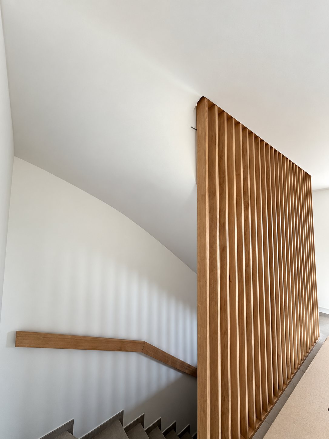 Une cage d'escalier aux murs blancs avec une main courante en bois et une cloison à claire-voie en lames de bois verticales.