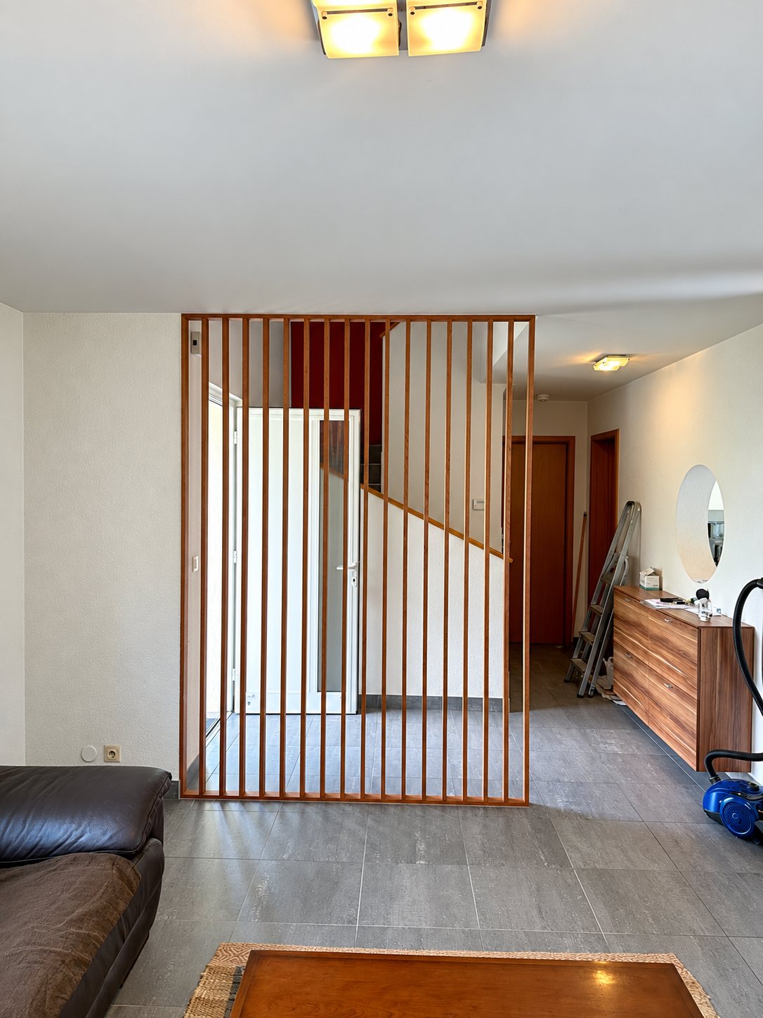 Une cloison claustra en lames de bois verticales sépare le salon de l'entrée, avec un escalier et une porte blanche visibles derrière.