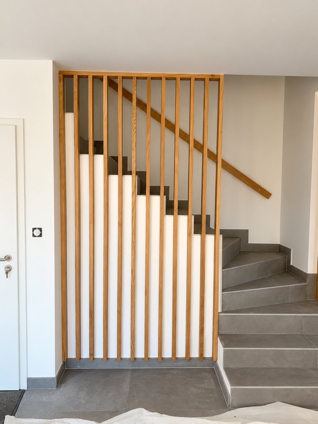 Un claustra en lames de bois clair verticales sépare l'entrée d'un escalier tournant aux marches carrelées grises, avec une main courante en bois.