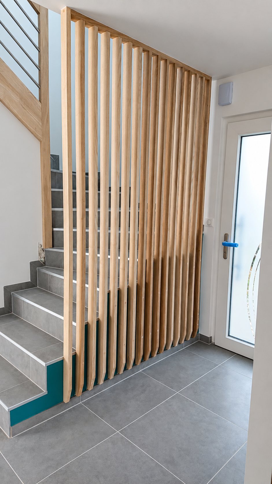 Une cloison claustra en lattes de bois clair verticales sépare l'entrée d'un escalier aux marches carrelées grises, près d'une porte vitrée blanche.