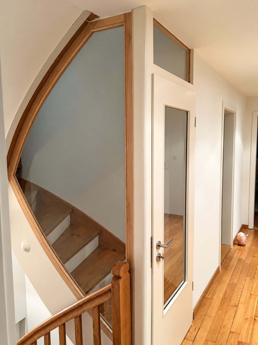 Dans un couloir lumineux, une cloison vitrée au cadre en bois clair épouse la courbe de l'escalier et comprend une porte blanche vitrée avec imposte.