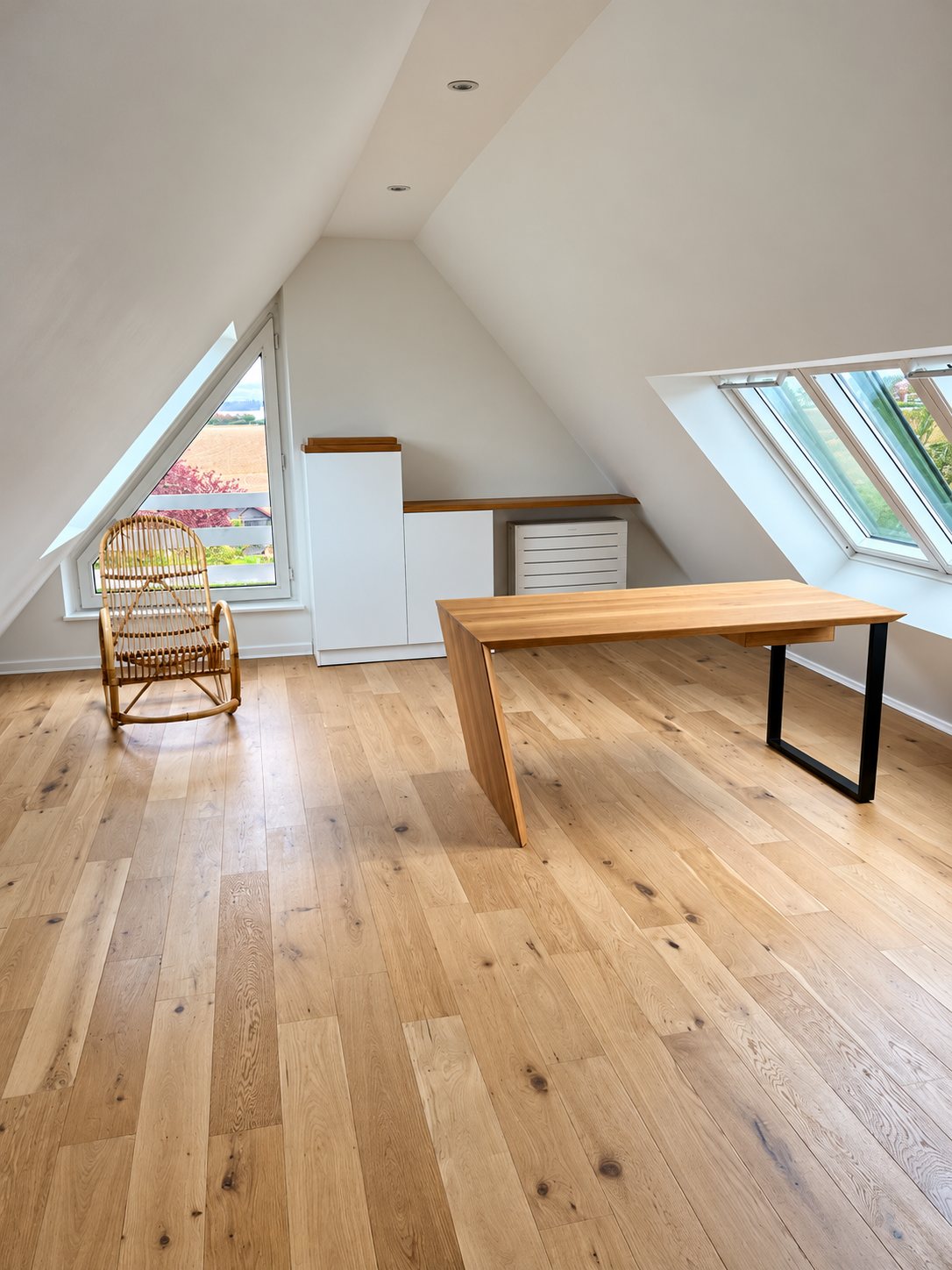 Pièce sous les combles au parquet de chêne clair, avec un bureau en bois à pied métallique noir, un meuble blanc sur mesure et un fauteuil à bascule en rotin.