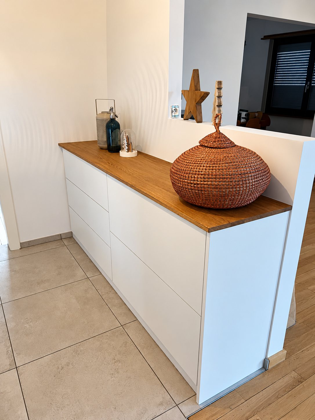 Commode blanche sur mesure à plateau en bois, ornée d'un panier tressé orange, d'une étoile en bois et d'objets décoratifs, installée contre une cloison dans une entrée carrelée.