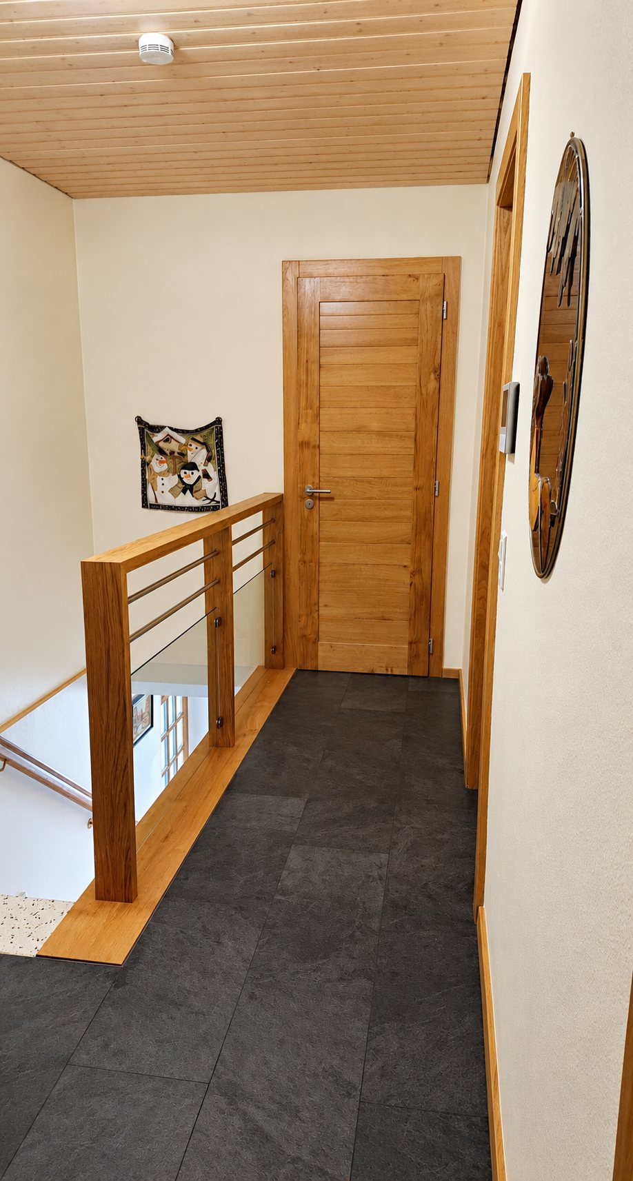 Un couloir lumineux avec un garde-corps en bois et métal, une porte en chêne et un sol en dalles sombres, au-dessus d'une cage d'escalier.