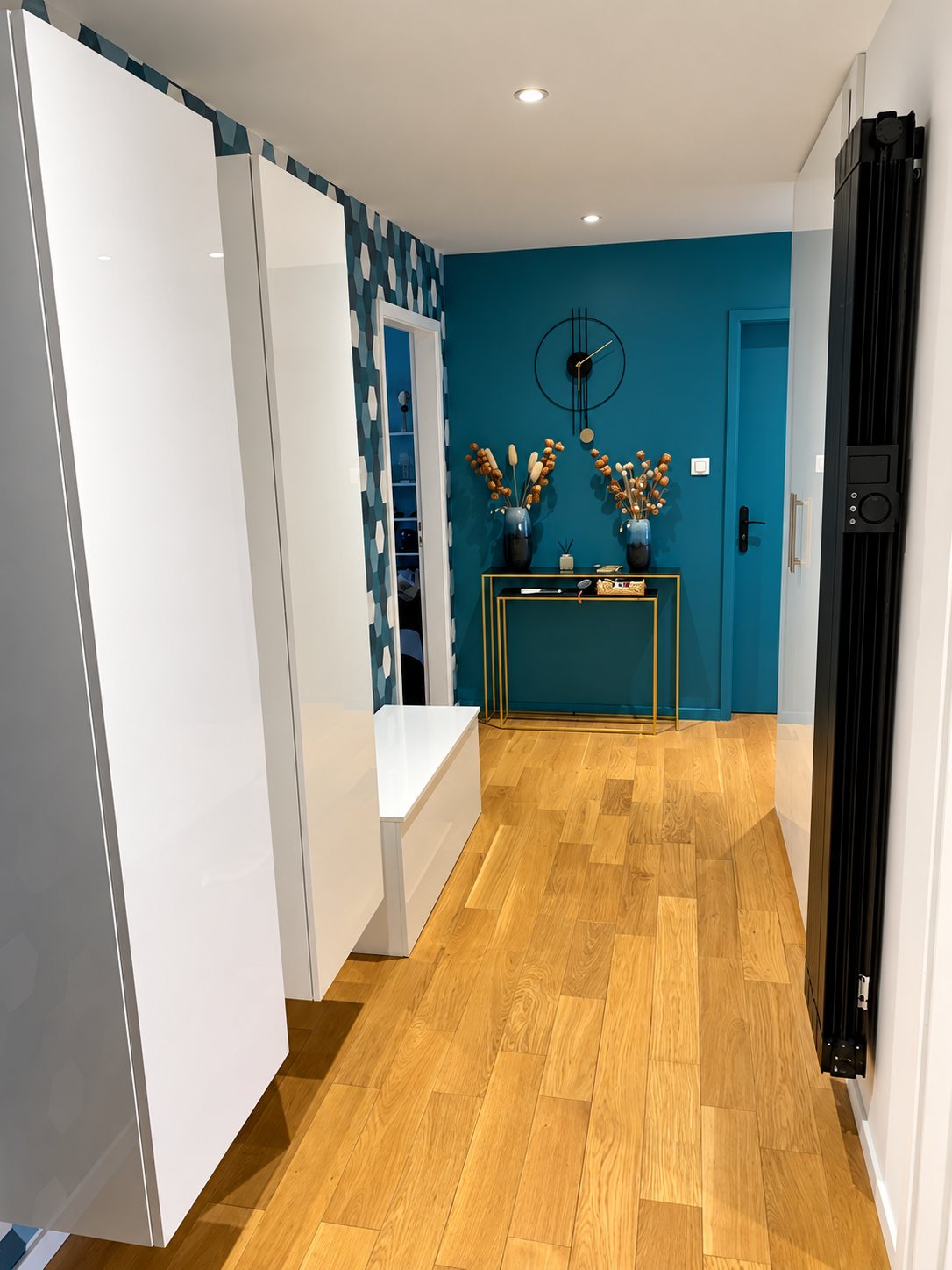 Un couloir moderne avec parquet en chêne clair, mur bleu canard, console dorée, horloge noire, meubles blancs laqués et radiateur noir vertical.