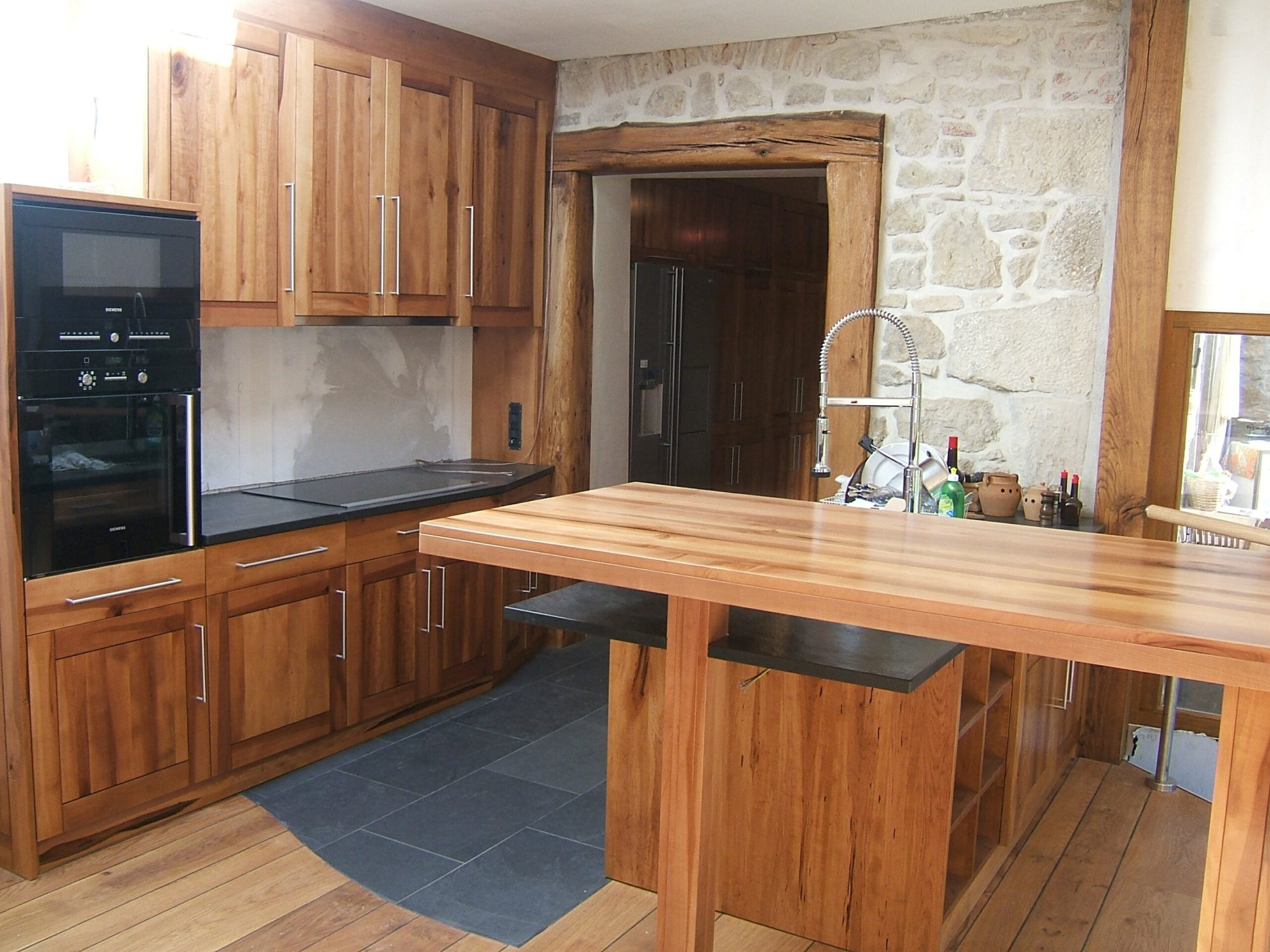 Cuisine sur mesure en chêne massif avec îlot central, plan de travail noir, four encastré et mur en pierres apparentes.