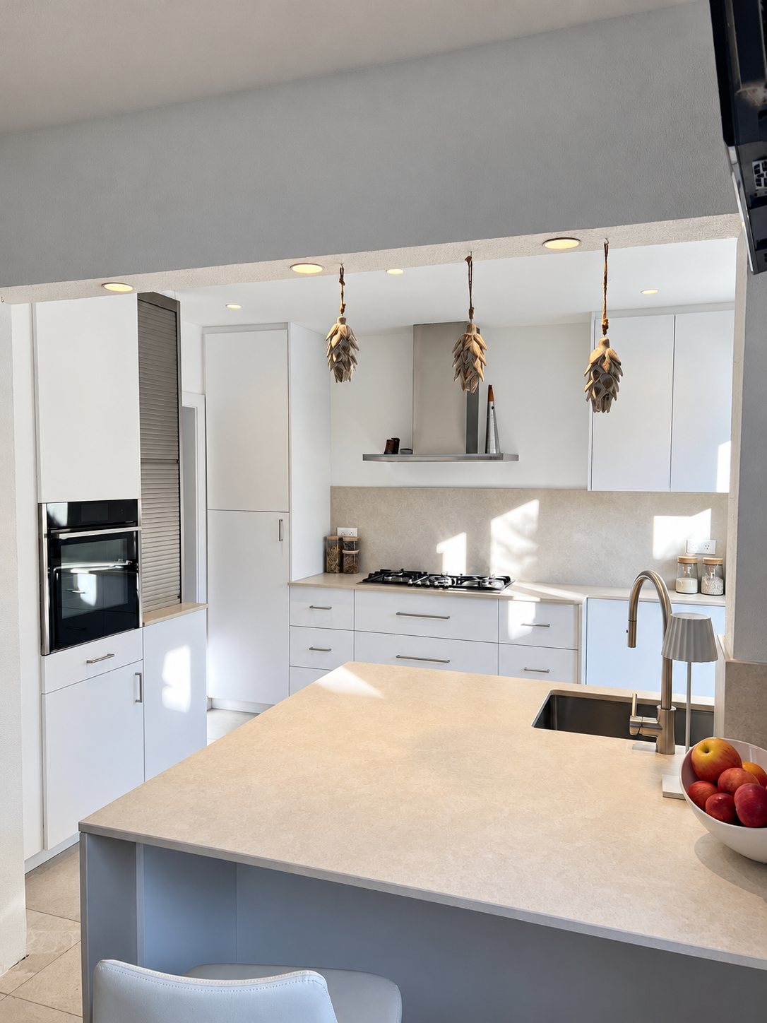 Cuisine contemporaine aux meubles blancs avec four encastré, plaque de cuisson, hotte inox et îlot central au plan de travail beige orné d'une coupe de pommes.
