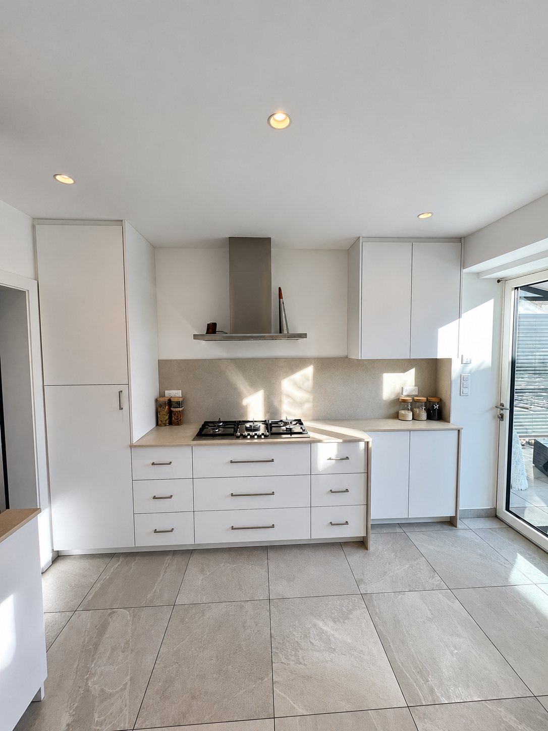 Cuisine moderne aux meubles blancs avec plaque de cuisson à gaz, hotte inox et grand carrelage gris, éclairée par le soleil.