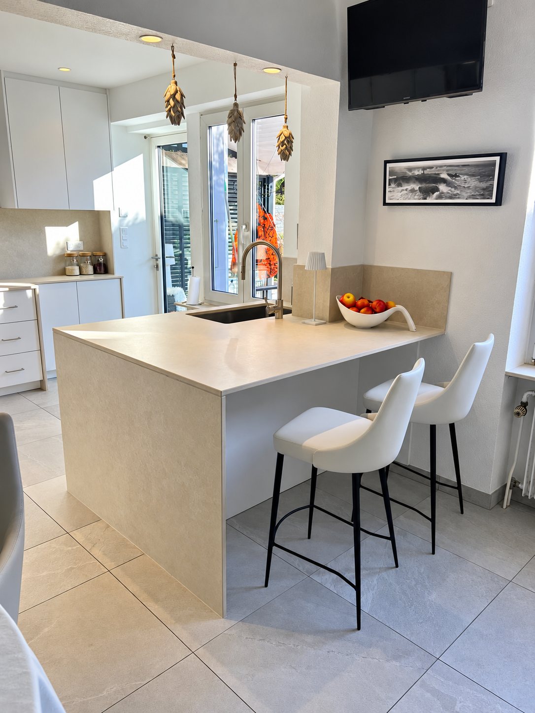 Cuisine lumineuse avec un îlot en pierre beige à chute, un évier, un robinet chromé, deux tabourets blancs et une corbeille de fruits.