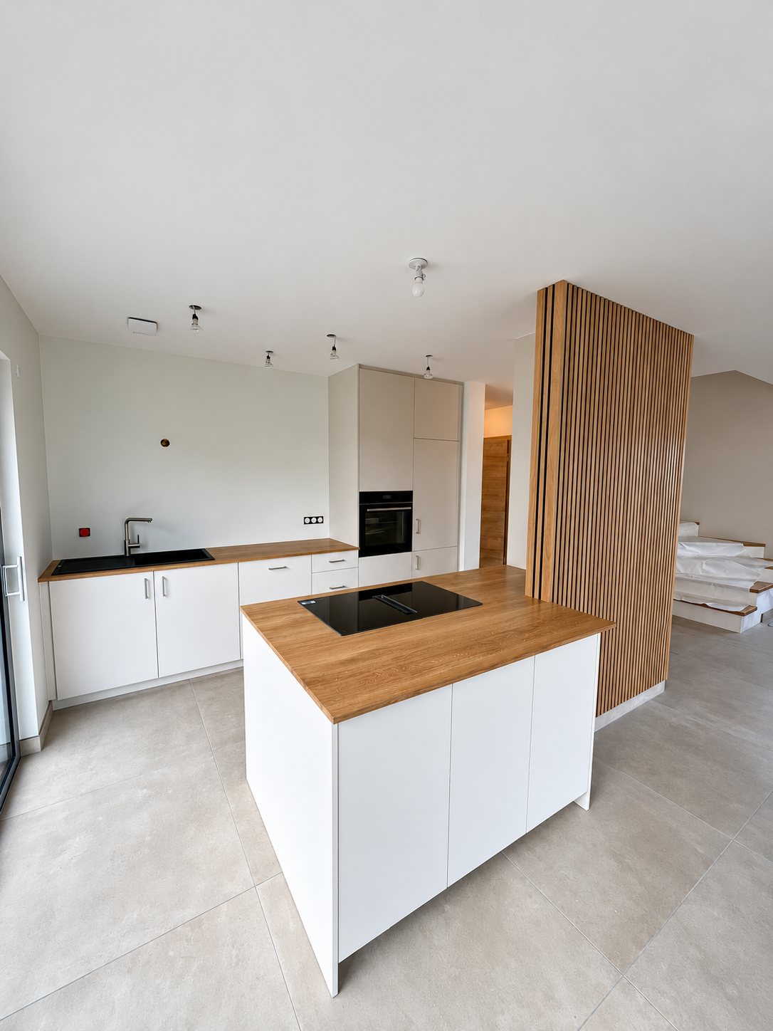 Cuisine neuve aux façades blanches avec un îlot central à plan de travail en chêne et plaque noire, à côté d'un panneau à claustra en bois.