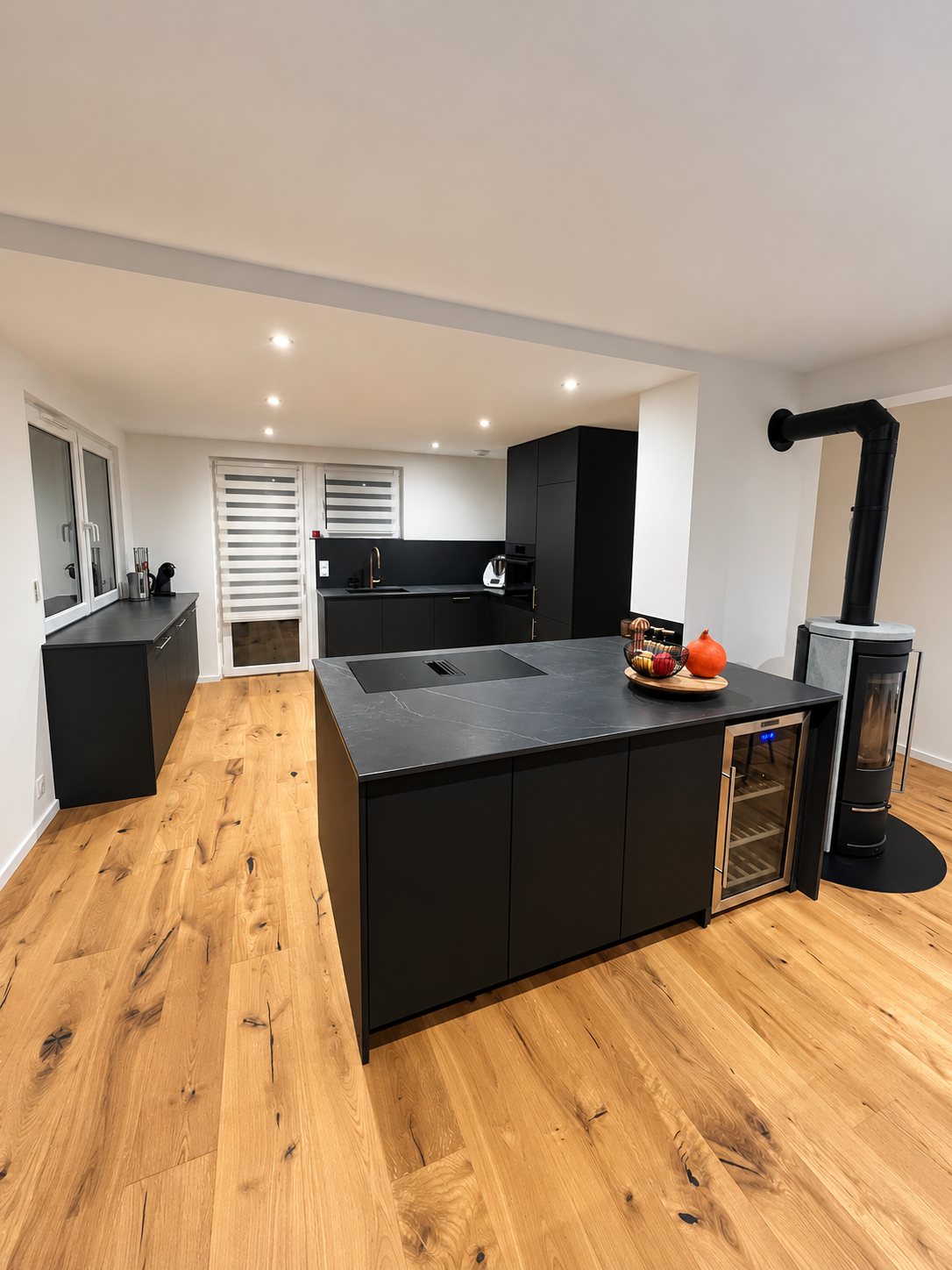 Cuisine moderne aux meubles noirs mats avec grand îlot central, cave à vin encastrée, parquet en chêne et poêle à bois dans la pièce.
