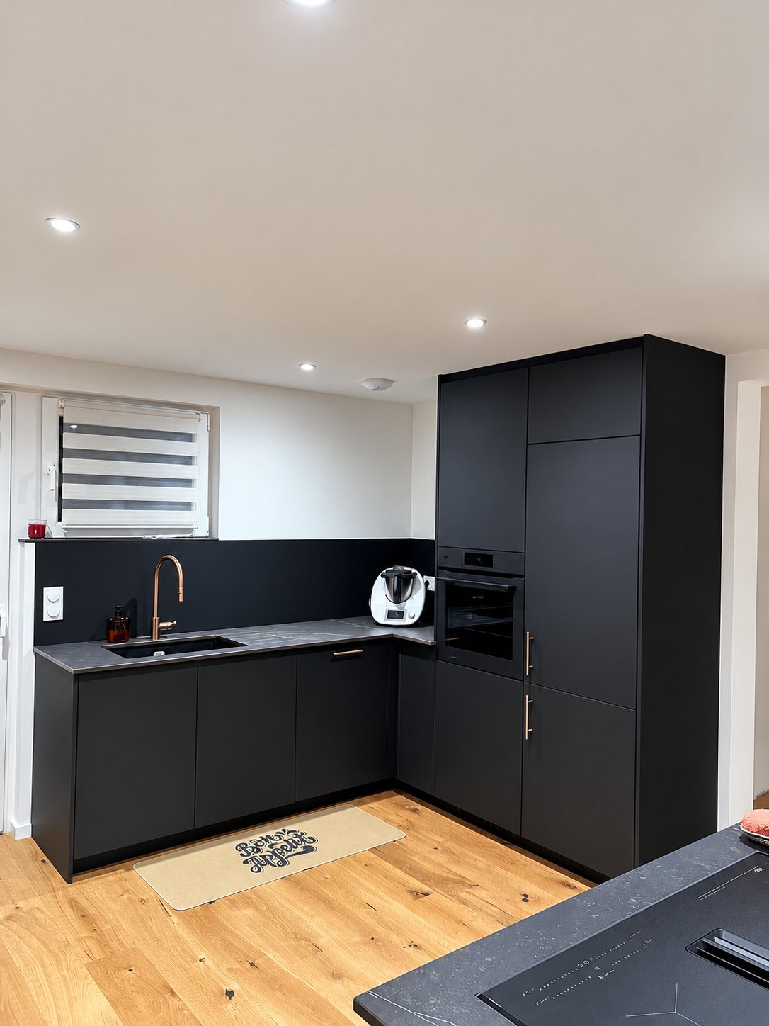 Cuisine équipée en angle aux façades noires mates, plan de travail sombre, robinet cuivré, four encastré et parquet clair, avec un tapis Bon Appétit au sol.