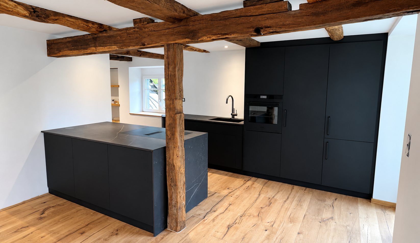 Cuisine noire mat avec îlot central et colonnes encastrées, sous des poutres en bois anciennes et sur un parquet en chêne clair.