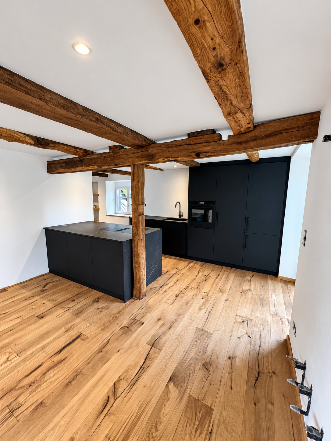 Une cuisine moderne noire mat avec îlot et colonnes, installée dans une pièce rénovée aux poutres en bois apparentes et au parquet en chêne.
