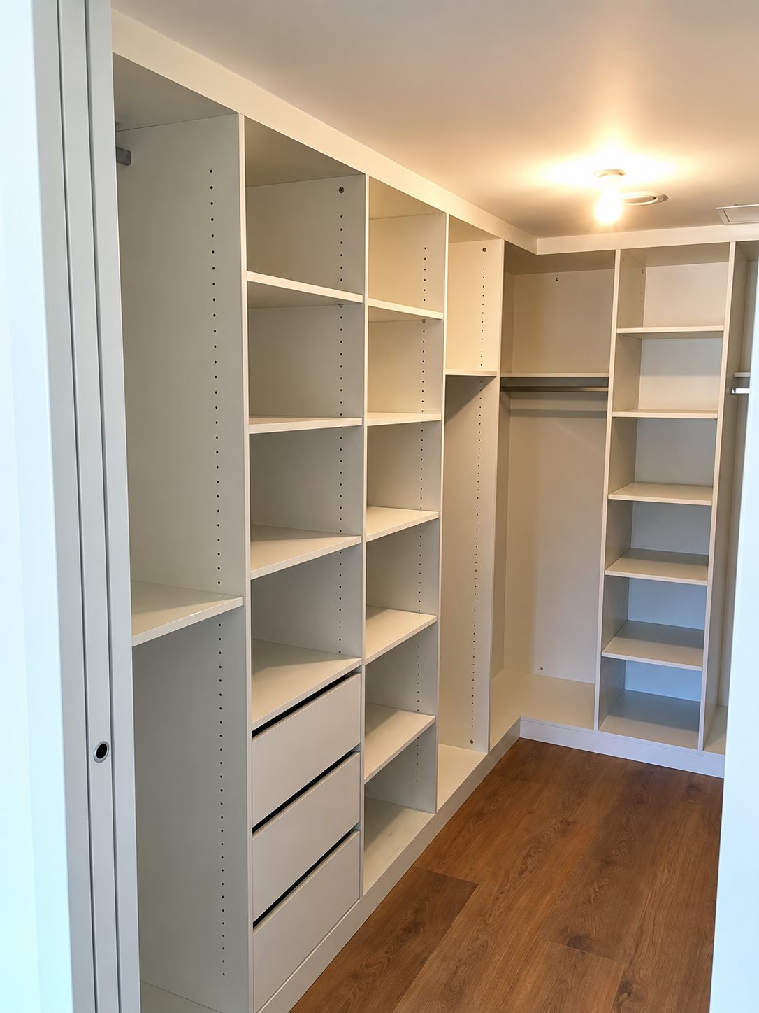 Un dressing d'angle vide aux étagères et tiroirs blancs, installé dans une pièce au parquet foncé.