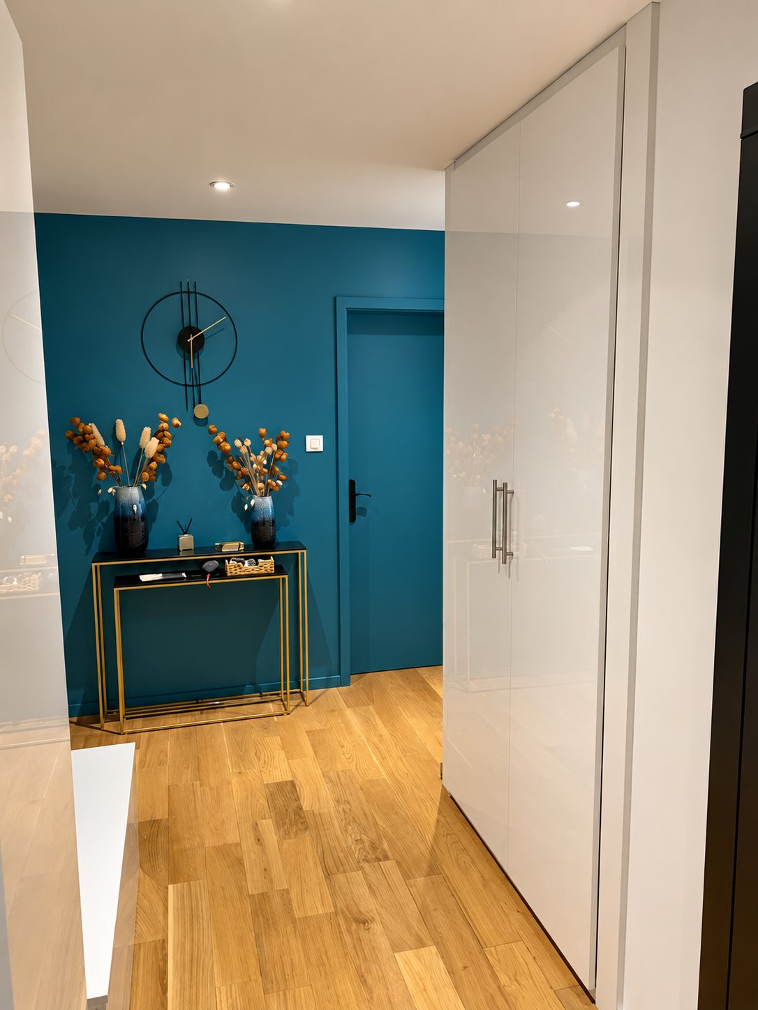 Entrée aménagée avec un mur bleu canard, une horloge noire, une console dorée ornée de vases de fleurs séchées, un parquet clair et un placard blanc laqué.