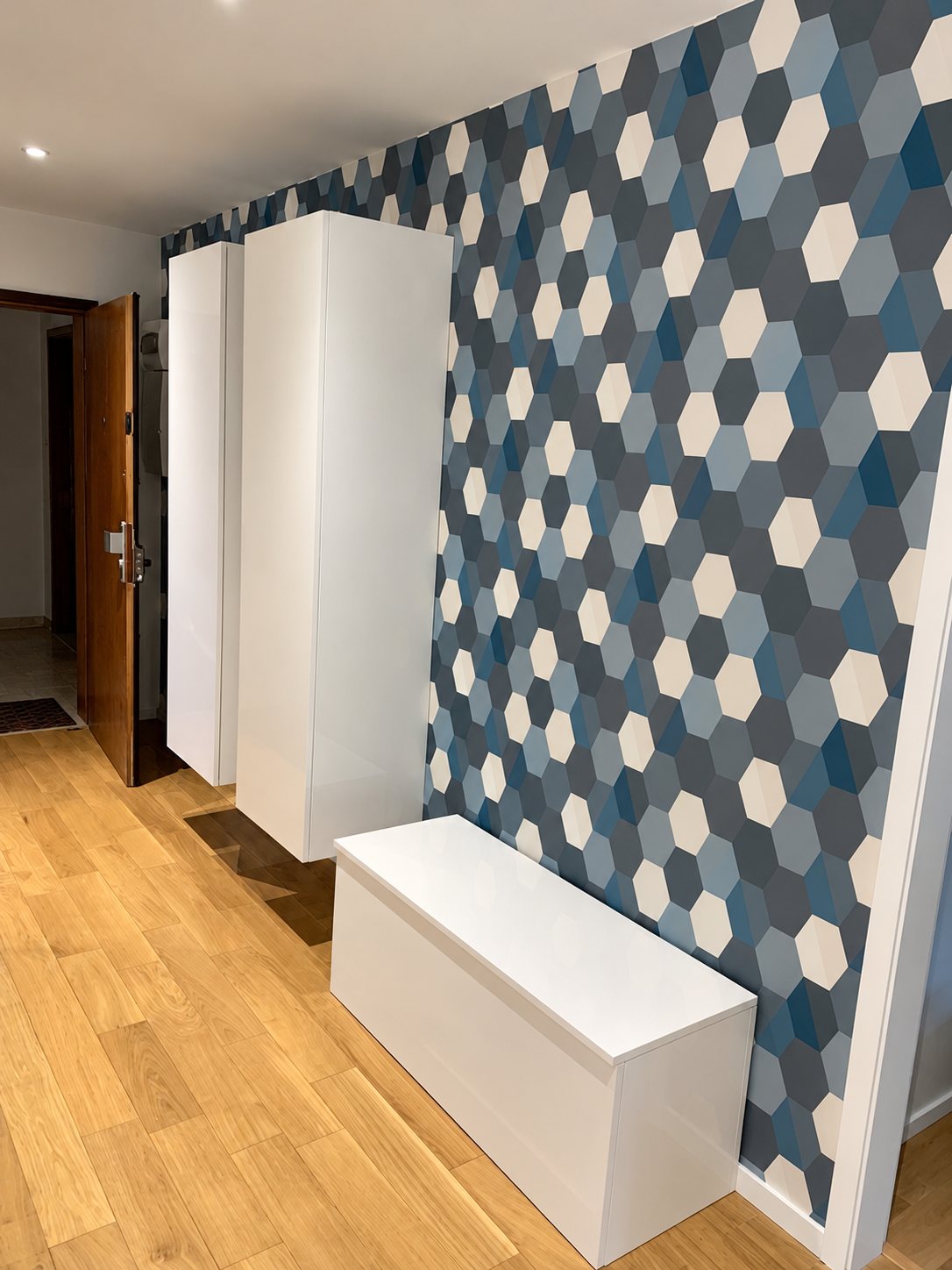 Entrée d'appartement avec un mur en papier peint à motifs hexagonaux bleus et gris, deux colonnes et un banc coffre blancs laqués sur un parquet en chêne.