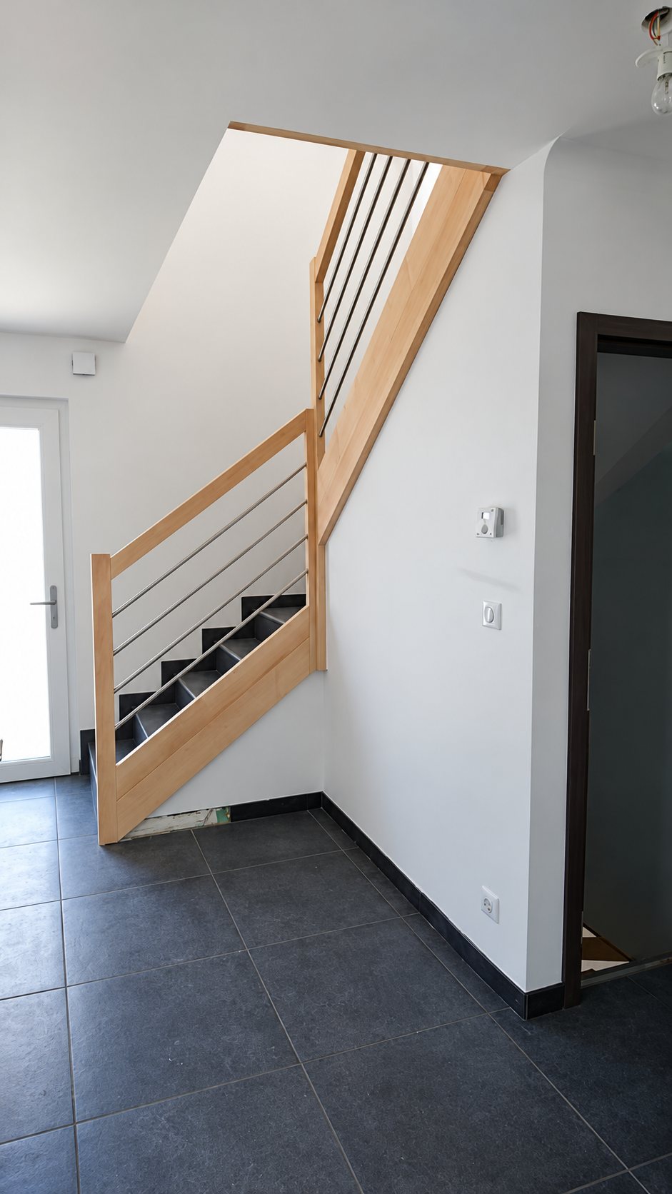 Un escalier tournant en bois clair avec rampe à barres métalliques horizontales et marches sombres, installé dans un intérieur aux murs blancs et au sol en carrelage gris anthracite.