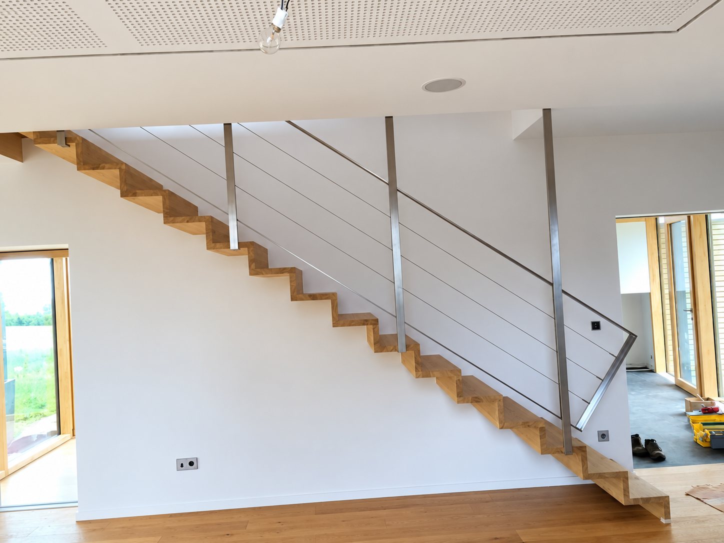 Un escalier droit en bois clair équipé d'un garde-corps en métal à câbles tendus, installé dans un intérieur moderne aux murs blancs.