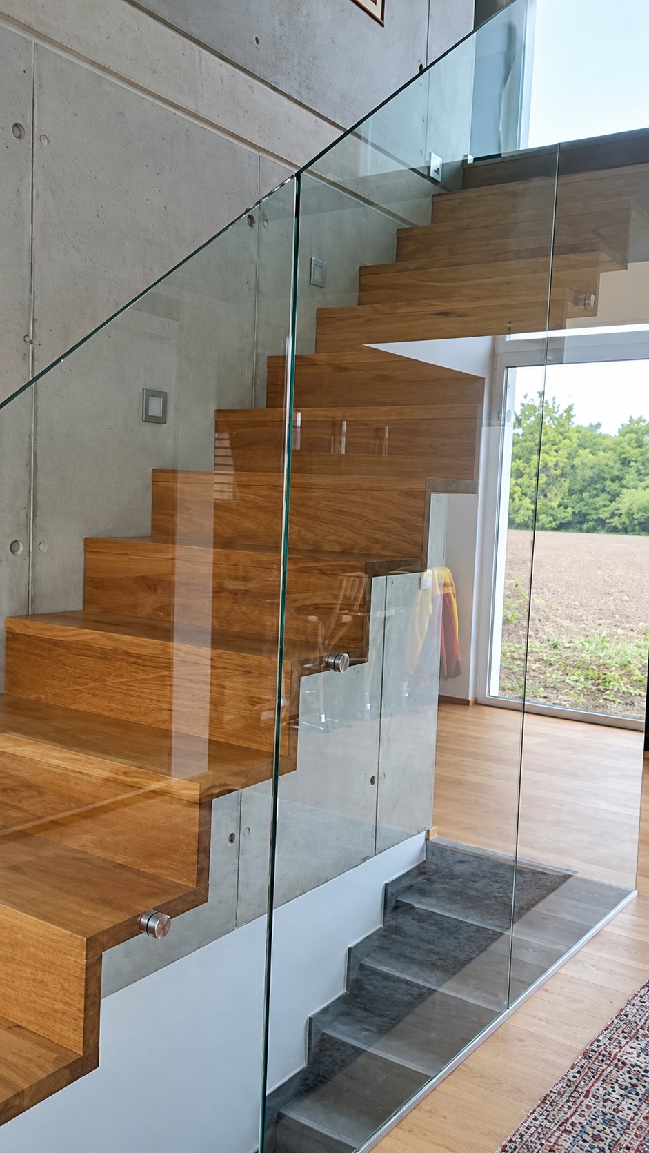 Un escalier moderne aux marches en bois massif protégé par un garde-corps en verre, dans un intérieur lumineux avec une grande baie vitrée.