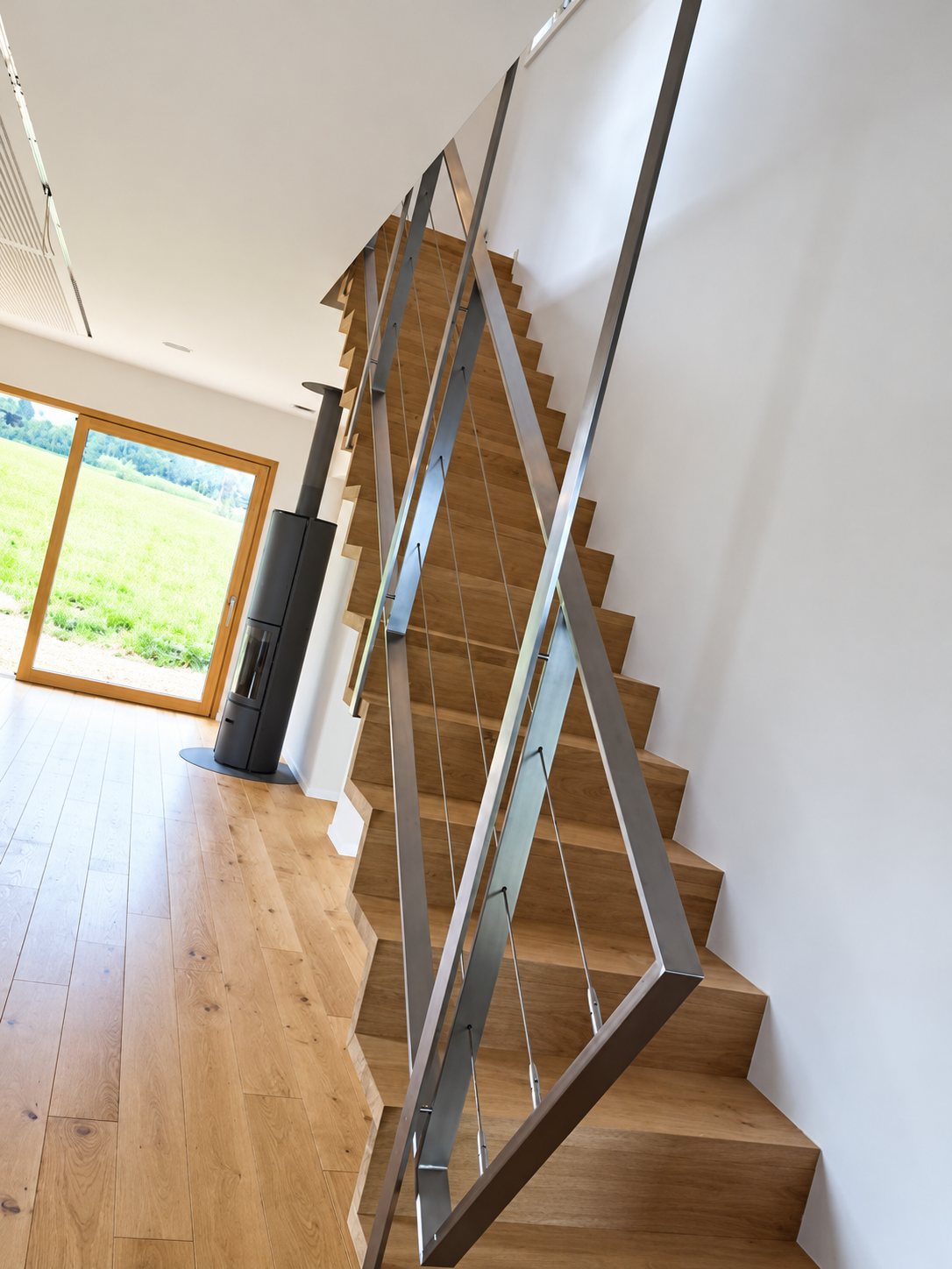 Un escalier droit en bois clair équipé d'une rampe en acier inoxydable à câbles tendus, dans une pièce lumineuse avec parquet et poêle à bois.