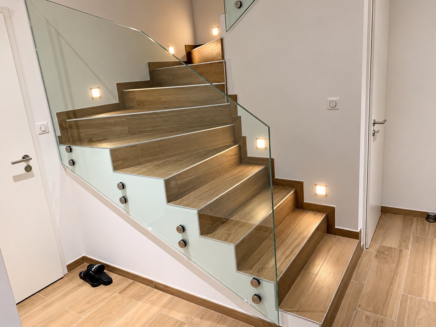 Escalier intérieur habillé de carrelage effet bois, équipé d'un garde-corps en verre fixé par des points inox et éclairé par des spots encastrés dans le mur.
