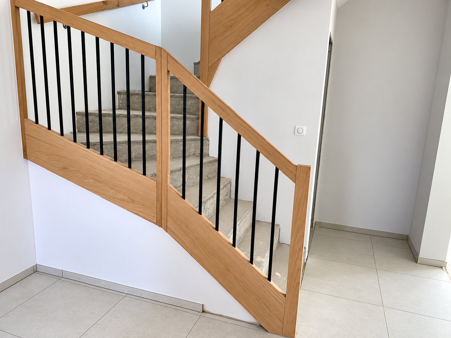 Escalier intérieur à garde-corps en chêne clair et barreaux noirs, avec marches en béton brut, dans une entrée aux murs blancs et au carrelage clair.