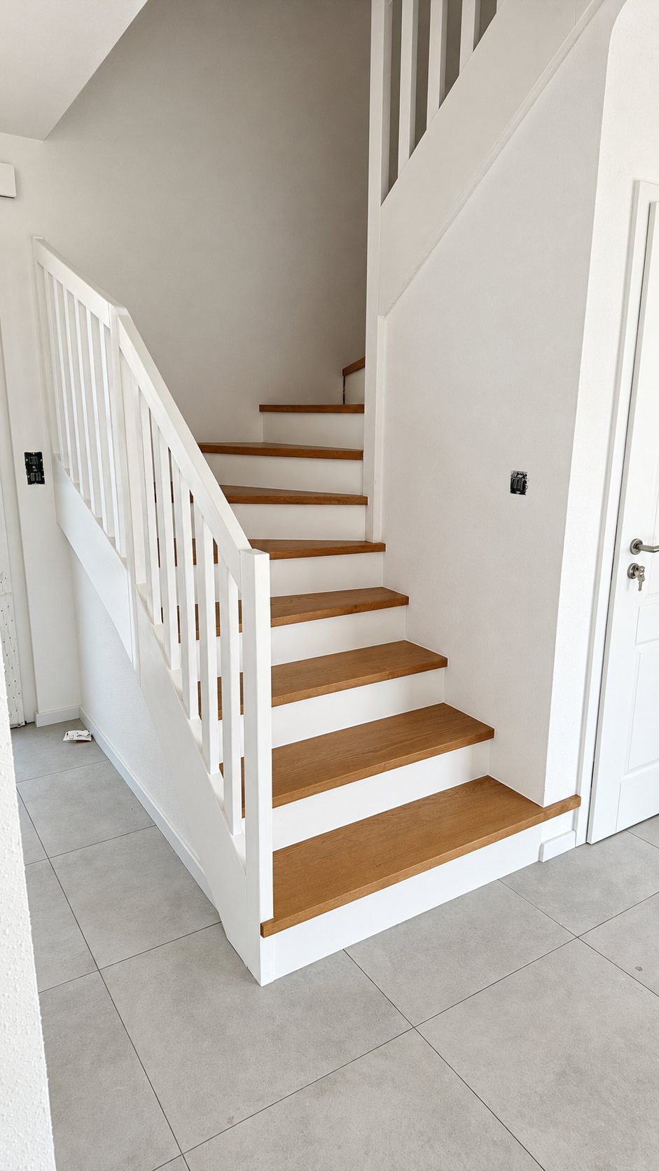 Un escalier tournant aux contremarches et à la balustrade blanches, avec des marches en bois clair, dans un hall au carrelage gris.