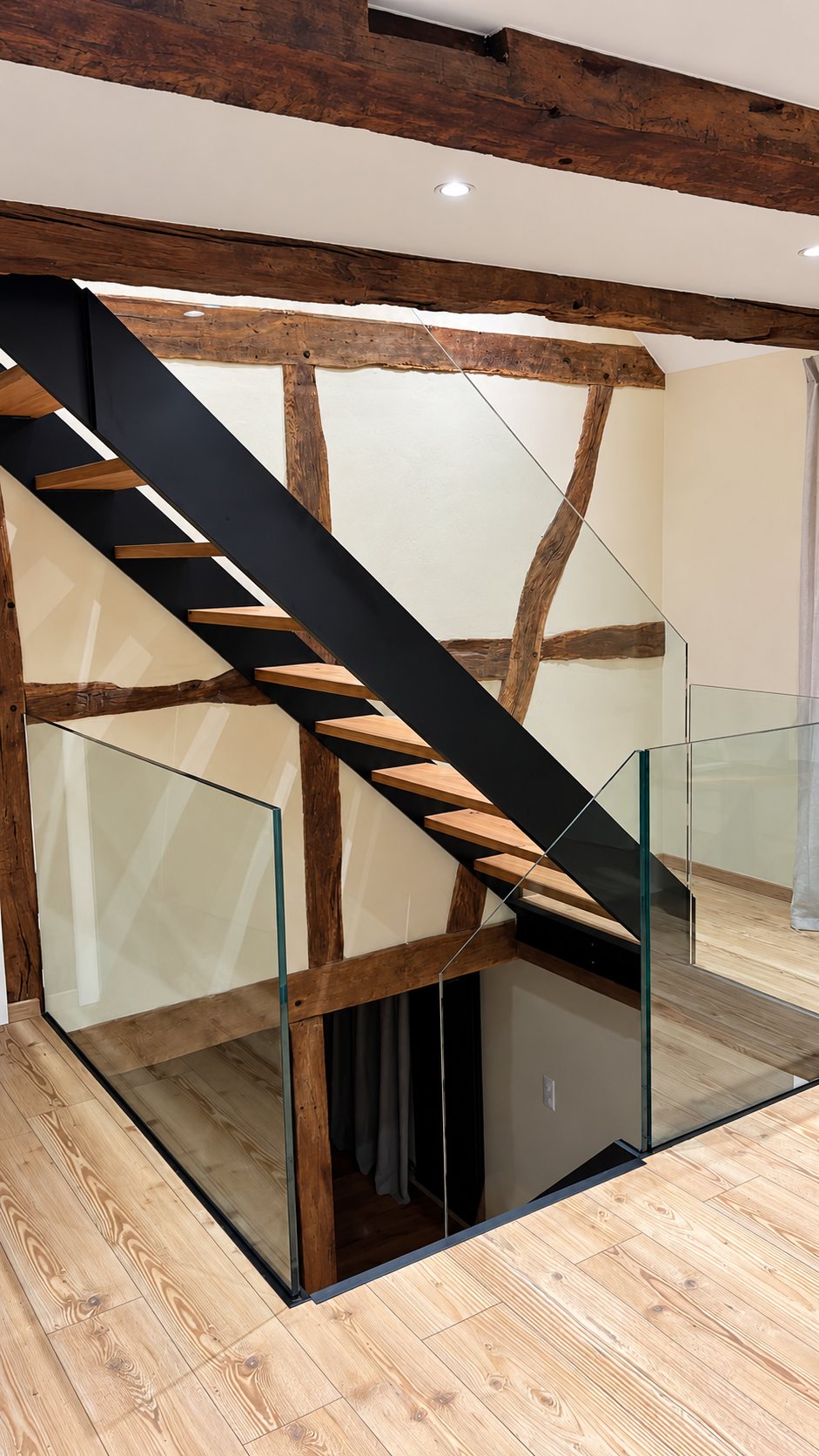 Un escalier au limon en acier noir et aux marches en bois, entouré de garde-corps en verre, dans une maison à colombages avec poutres anciennes et parquet clair.