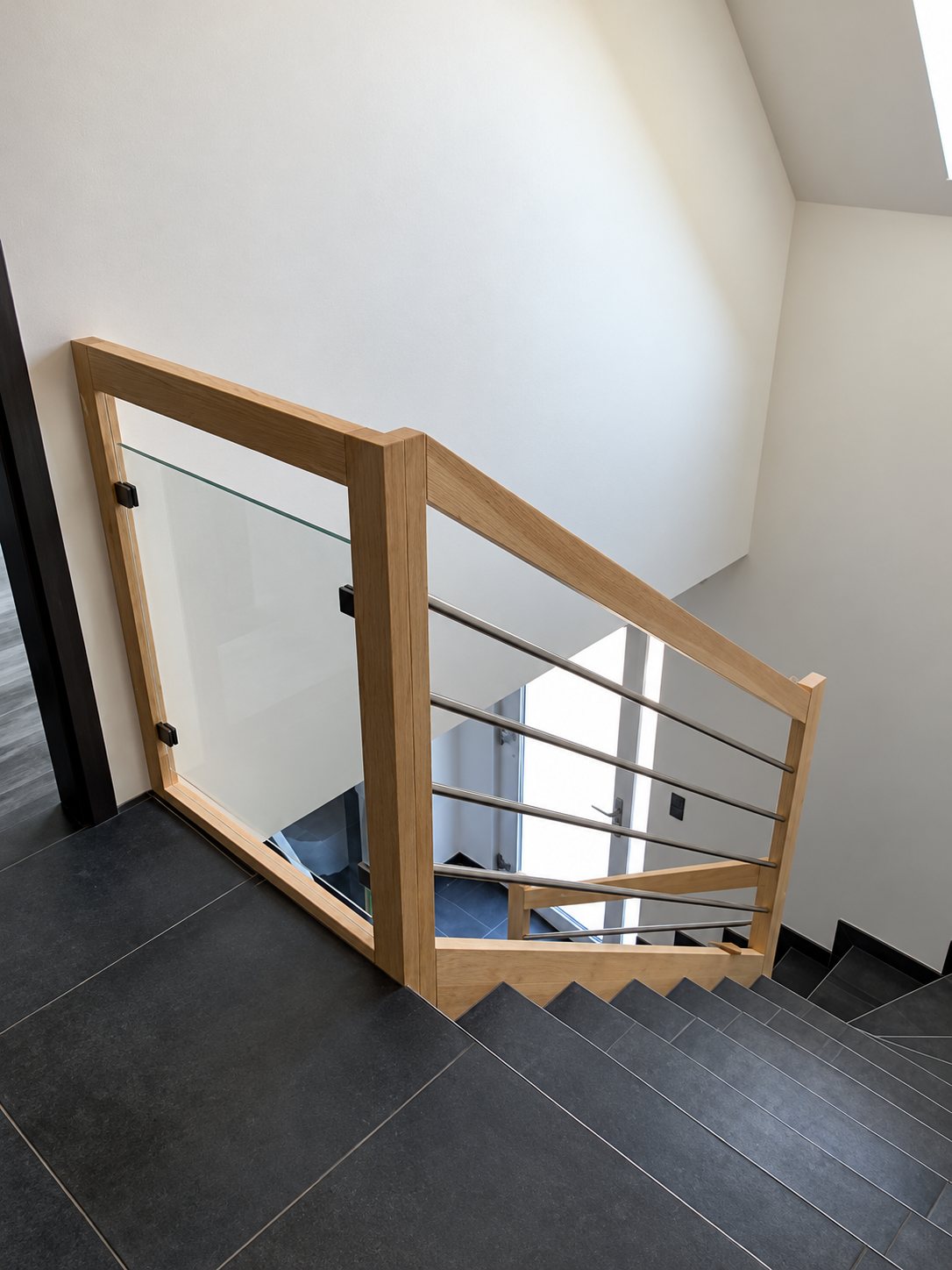 Vue plongeante sur un escalier aux marches sombres équipé d'une rampe en bois clair avec panneau de verre et câbles métalliques.