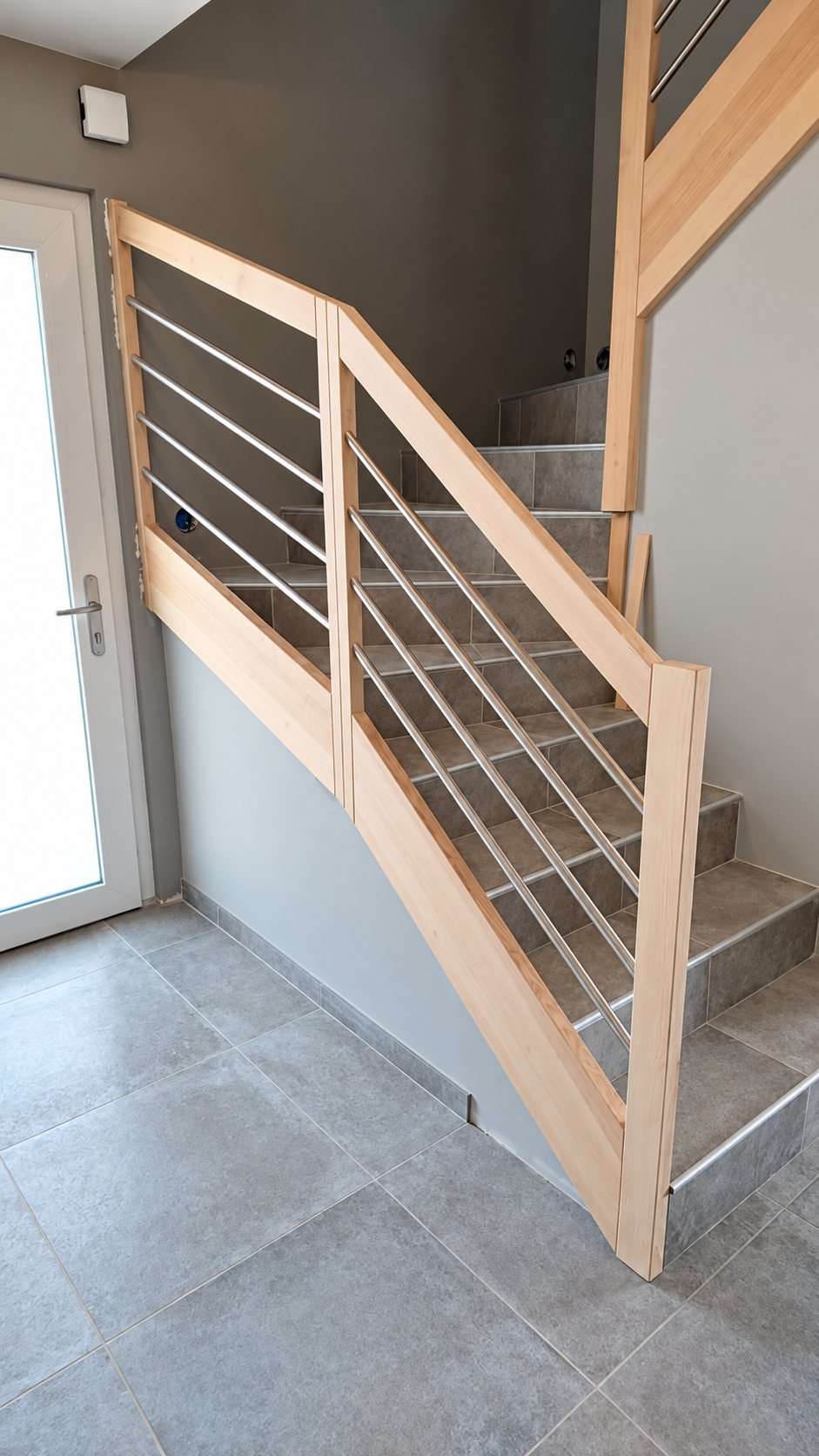 Escalier intérieur aux marches carrelées gris, équipé d'une rampe en bois clair avec barres métalliques horizontales, près d'une porte blanche vitrée.