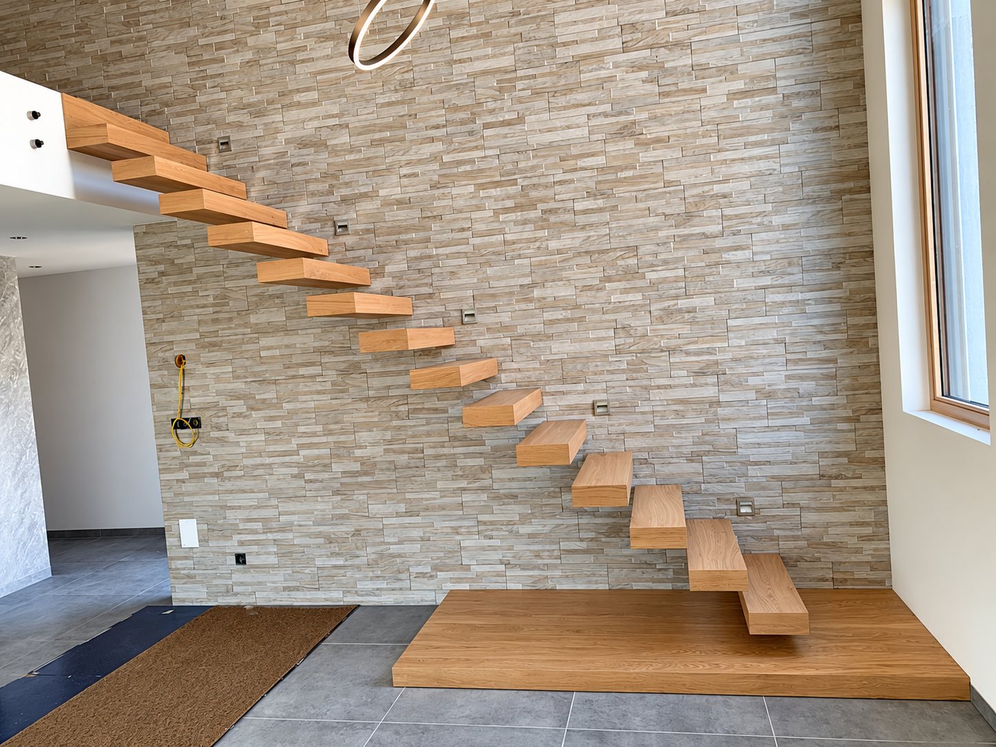 Escalier suspendu aux marches en bois de chêne fixées à un mur en parement de pierre beige, dans un intérieur moderne au carrelage gris.
