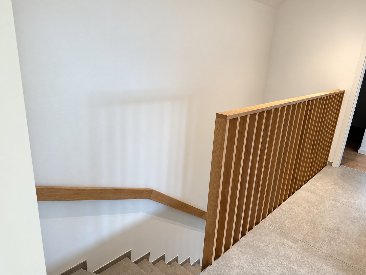 Garde-corps en chêne à lames verticales et main courante en bois sur un palier carrelé, au-dessus d'un escalier aux marches grises.