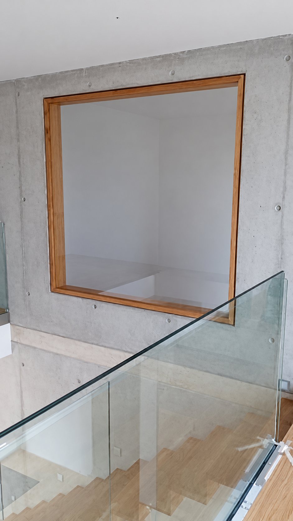 Un garde-corps en verre longe un escalier en bois, devant un mur en béton brut percé d'une grande fenêtre carrée à cadre en bois.