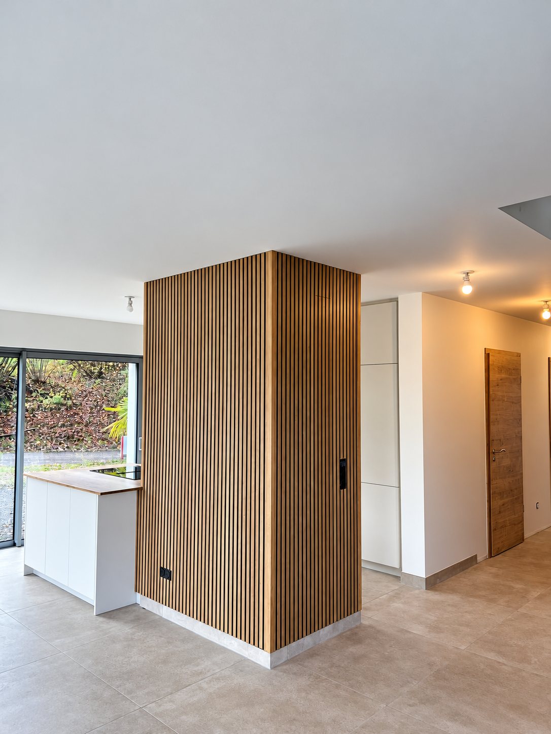 Un pan de mur habillé de tasseaux de bois verticaux dans un salon ouvert et lumineux, avec cuisine blanche, carrelage clair et porte en bois.