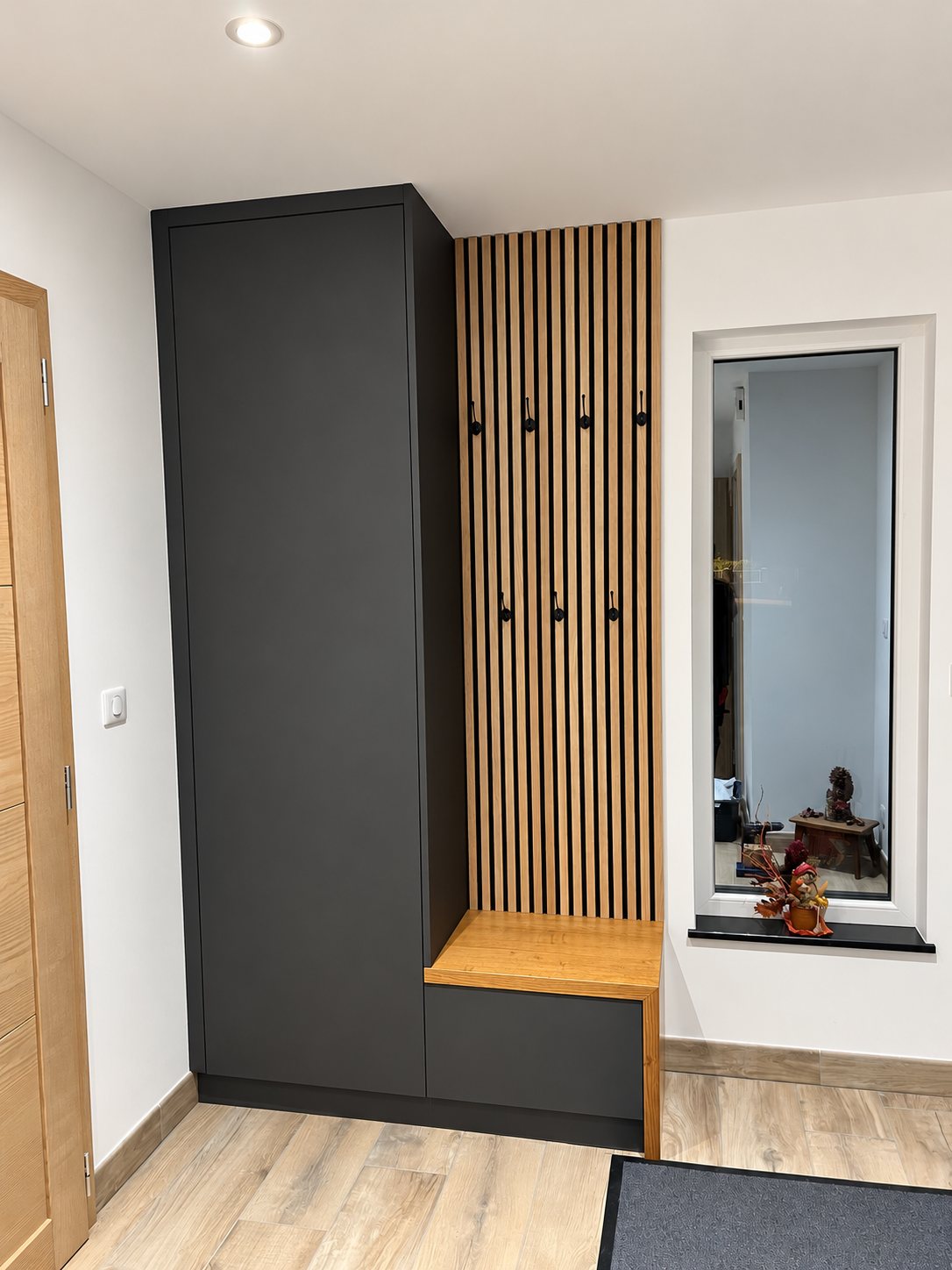 Meuble d'entrée sur mesure composé d'une armoire anthracite, d'un banc en bois et d'un panneau de tasseaux avec patères noires, à côté d'une fenêtre intérieure.
