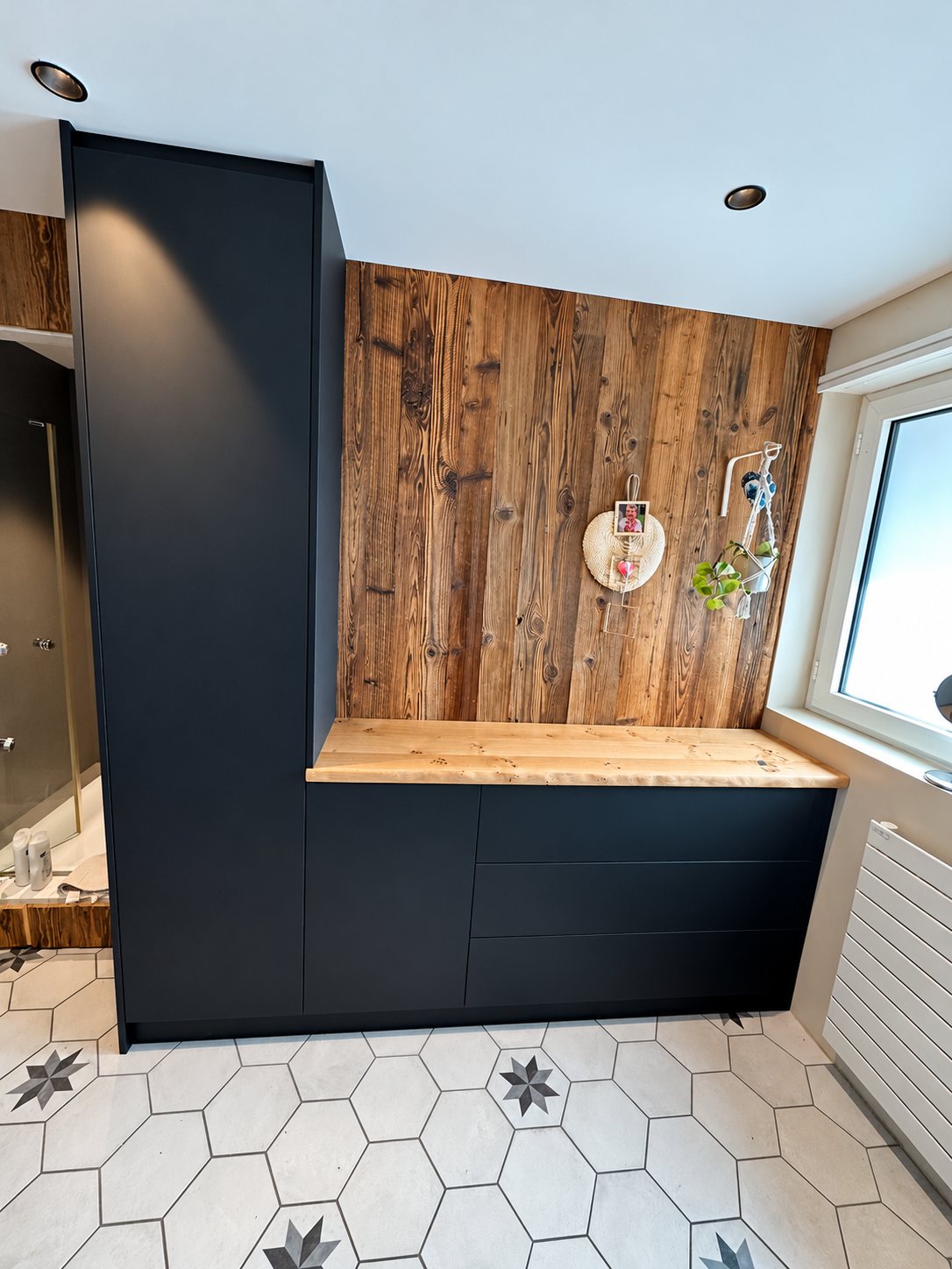Meuble sur mesure noir mat avec plan de travail en bois brut et mur en vieux bois, dans une salle de bain au carrelage hexagonal à motifs d'étoiles.