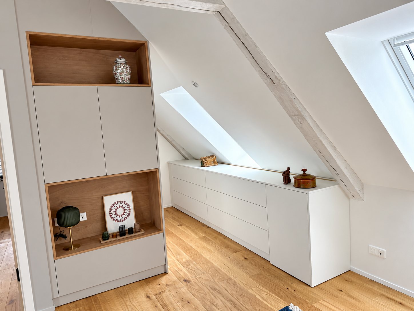 Dans des combles aux poutres apparentes, une armoire beige à niches en chêne et une commode basse blanche sur mesure sont installées sur un parquet clair.