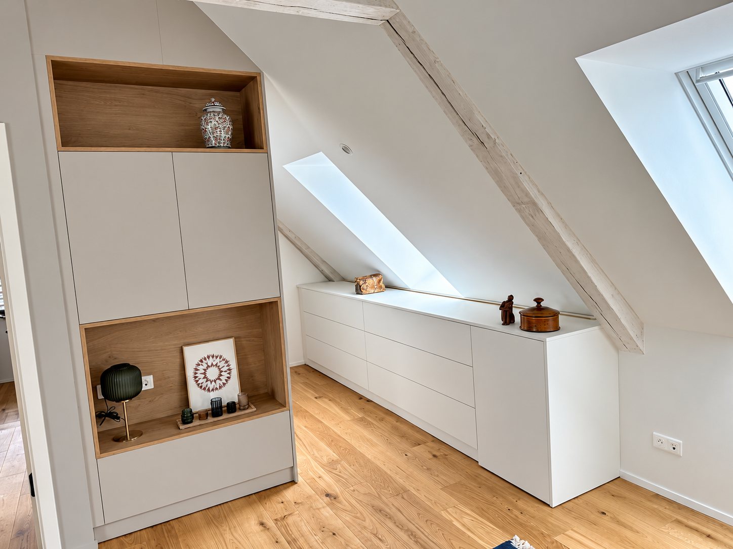Dans des combles lumineux, une bibliothèque blanche et chêne et une longue commode basse sous la pente du toit, avec parquet clair et fenêtres de toit.