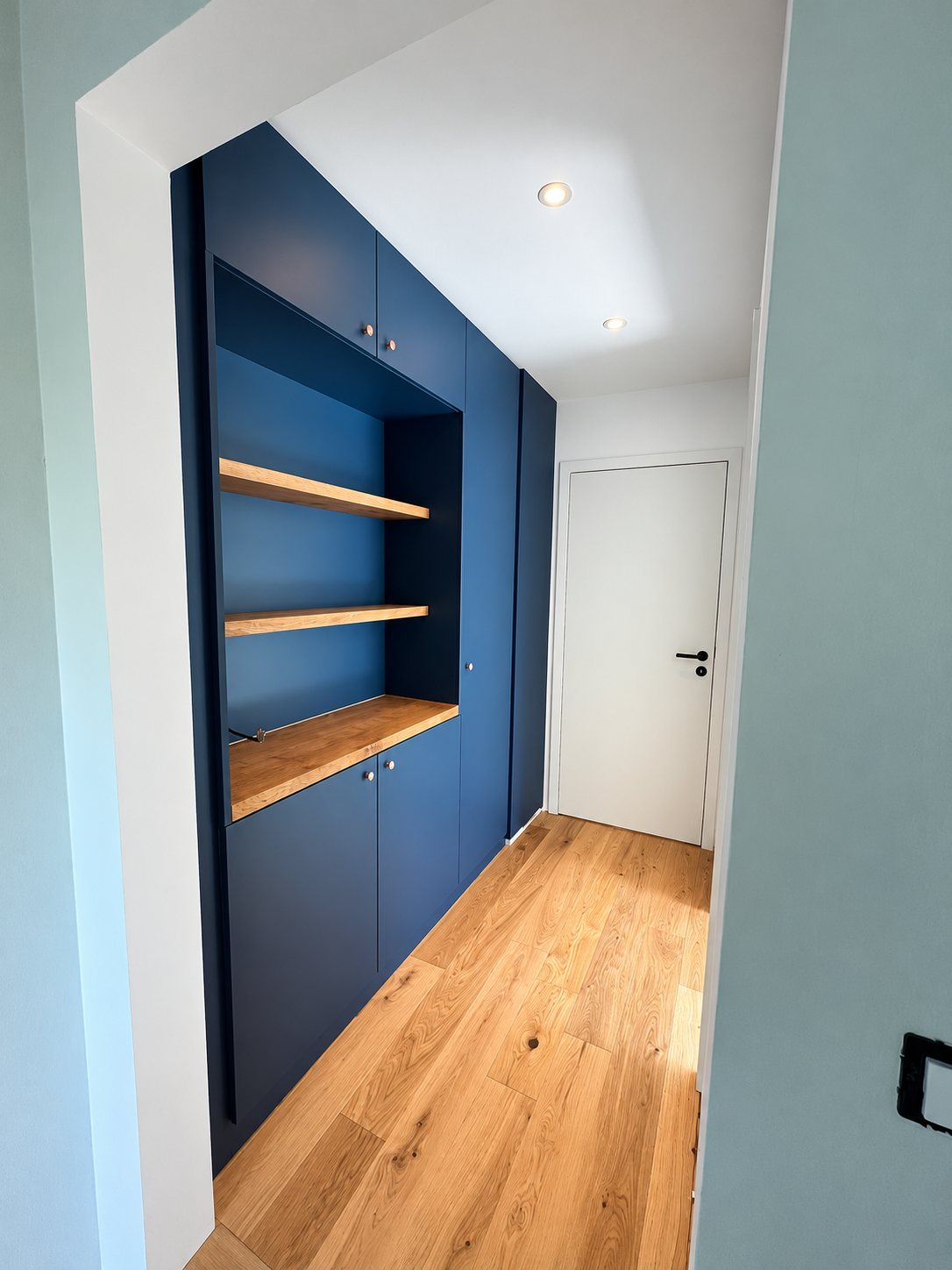 Un placard encastré bleu foncé avec des étagères en chêne clair longe un couloir au parquet en bois, devant une porte blanche.