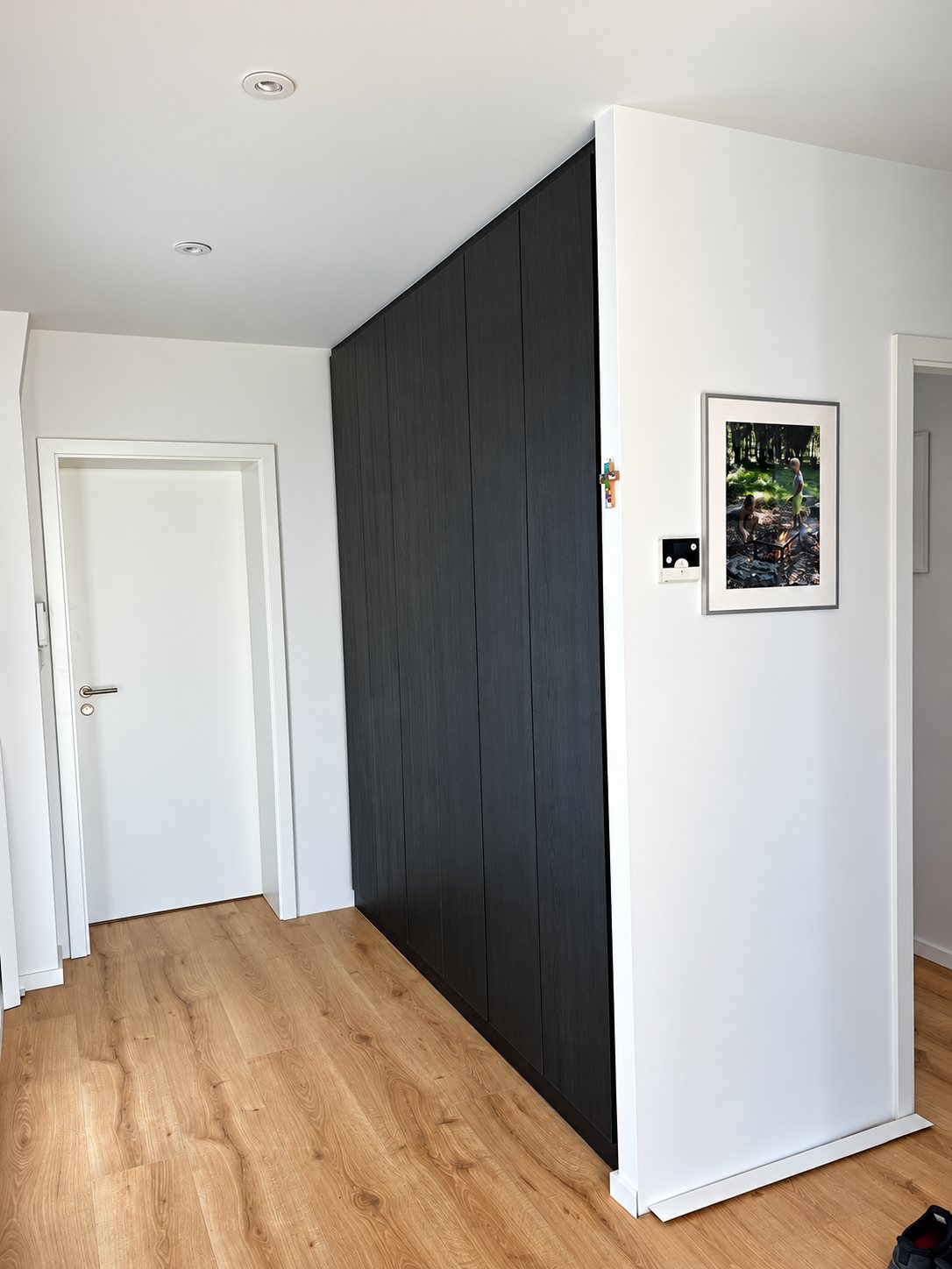 Un grand placard sur mesure en bois sombre, du sol au plafond, installé dans un couloir blanc avec parquet clair et un cadre au mur.