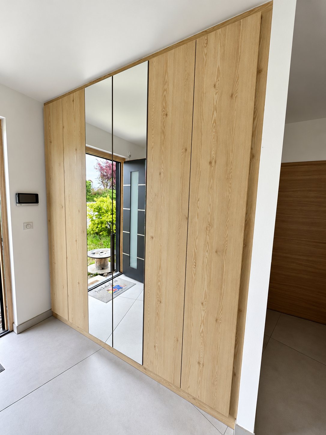Un grand placard sur mesure en bois clair avec deux portes miroir est installé dans une entrée lumineuse au sol en carrelage beige.
