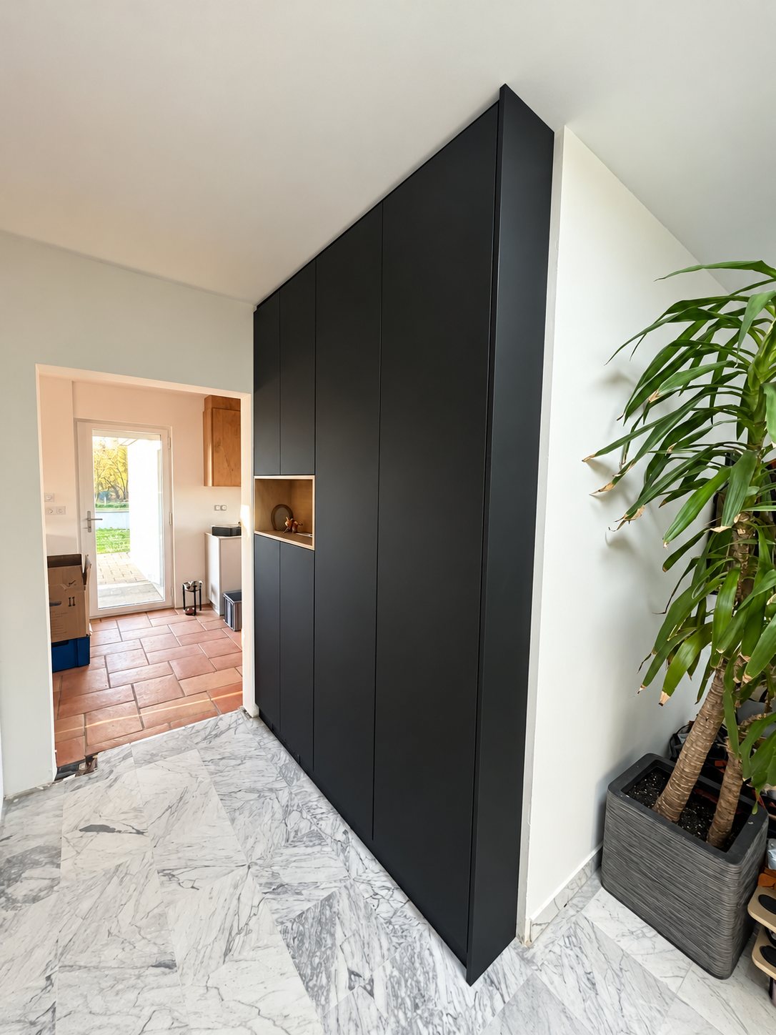 Un grand placard noir mat avec une niche en bois, installé dans une entrée au sol de marbre gris, à côté d'un yucca en pot.