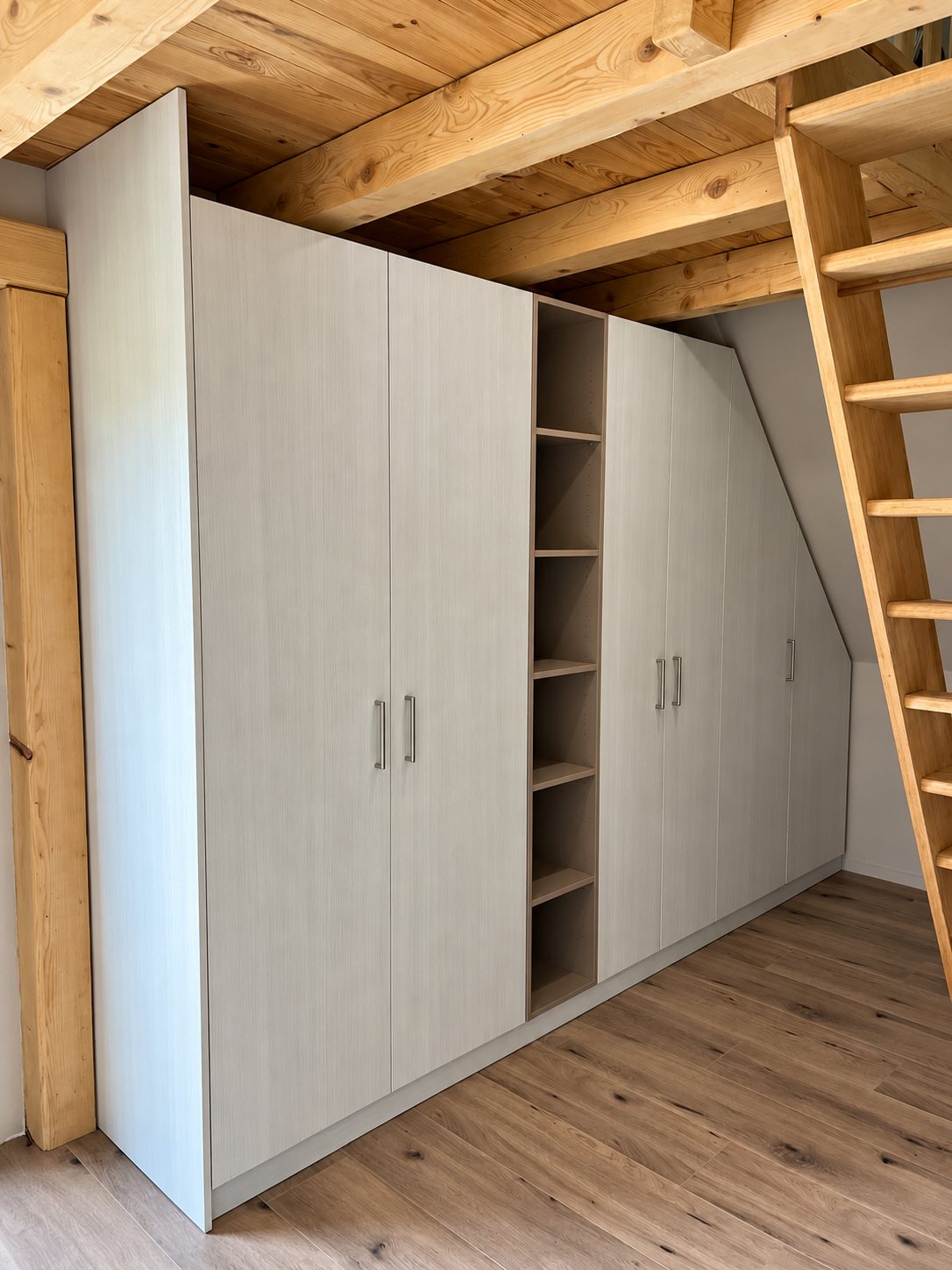 Un grand placard sur mesure gris clair avec une colonne d'étagères ouvertes est installé sous la pente d'un toit, près d'une échelle en bois et de poutres apparentes.