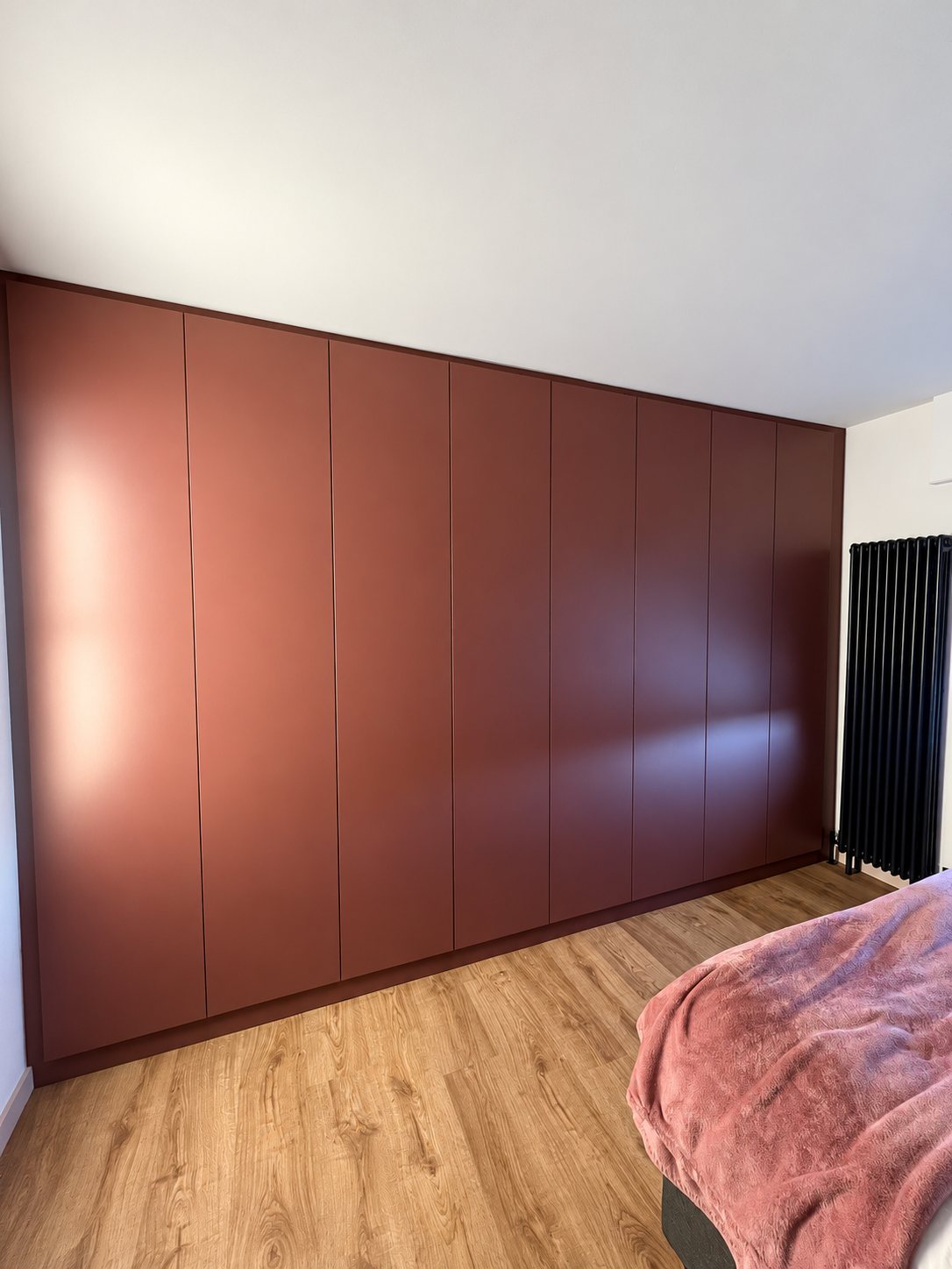 Une longue armoire murale sur mesure aux portes lisses terracotta occupe le mur d'une chambre au parquet clair, à côté d'un radiateur noir vertical et d'un lit avec plaid rose.