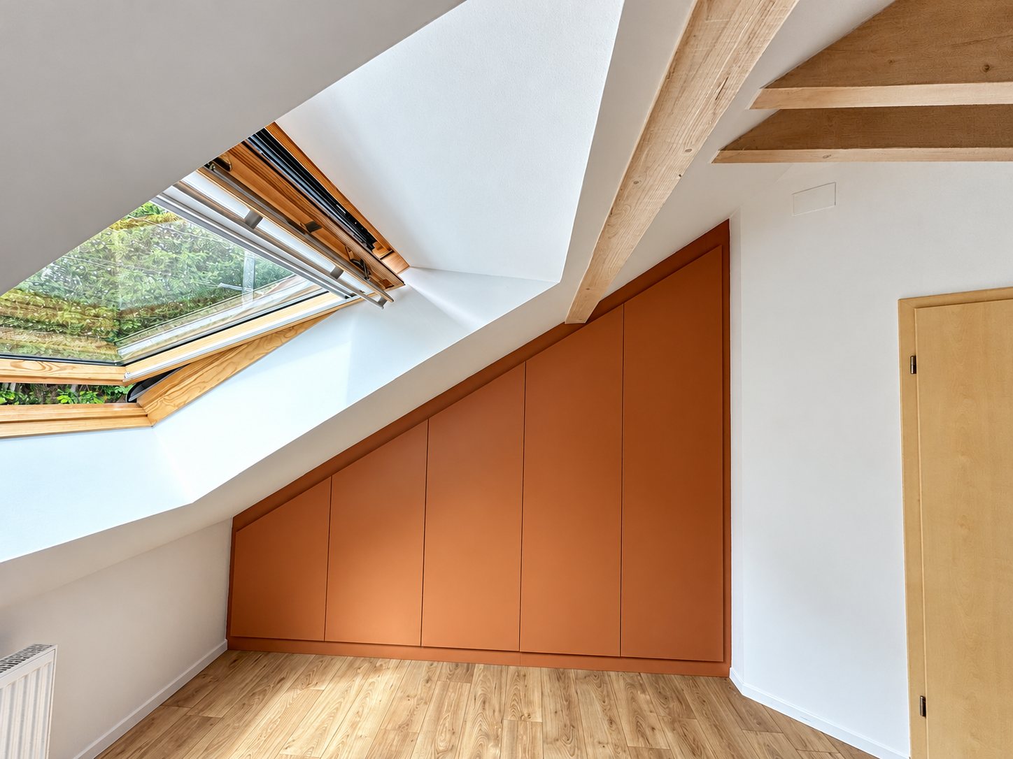 Sous un toit en pente, un placard sur mesure aux portes terracotta épouse la forme du mur, à côté d'une fenêtre de toit et d'une poutre en bois clair.